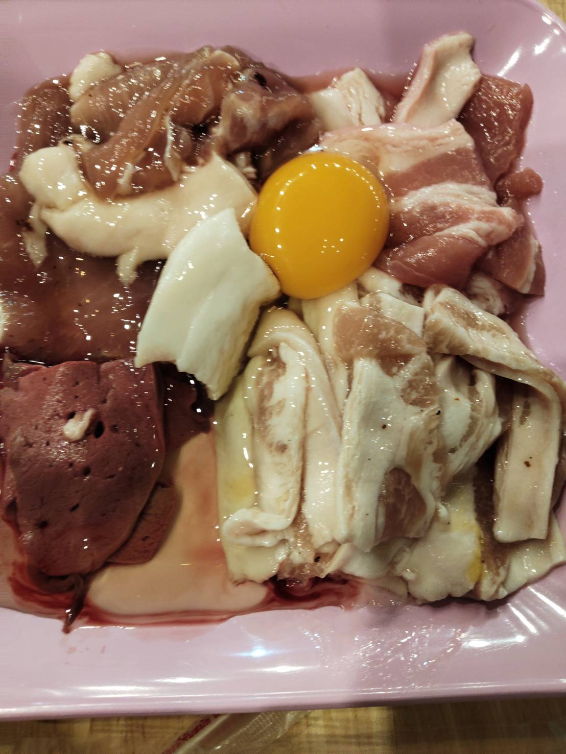 จานเนื้อสัตว์ดิบหลากชนิดพร้อมไข่แดงบนจานสีชมพู สำหรับปิ้งย่างหรือชาบู มีเนื้อหมู ตับ และมันหมู