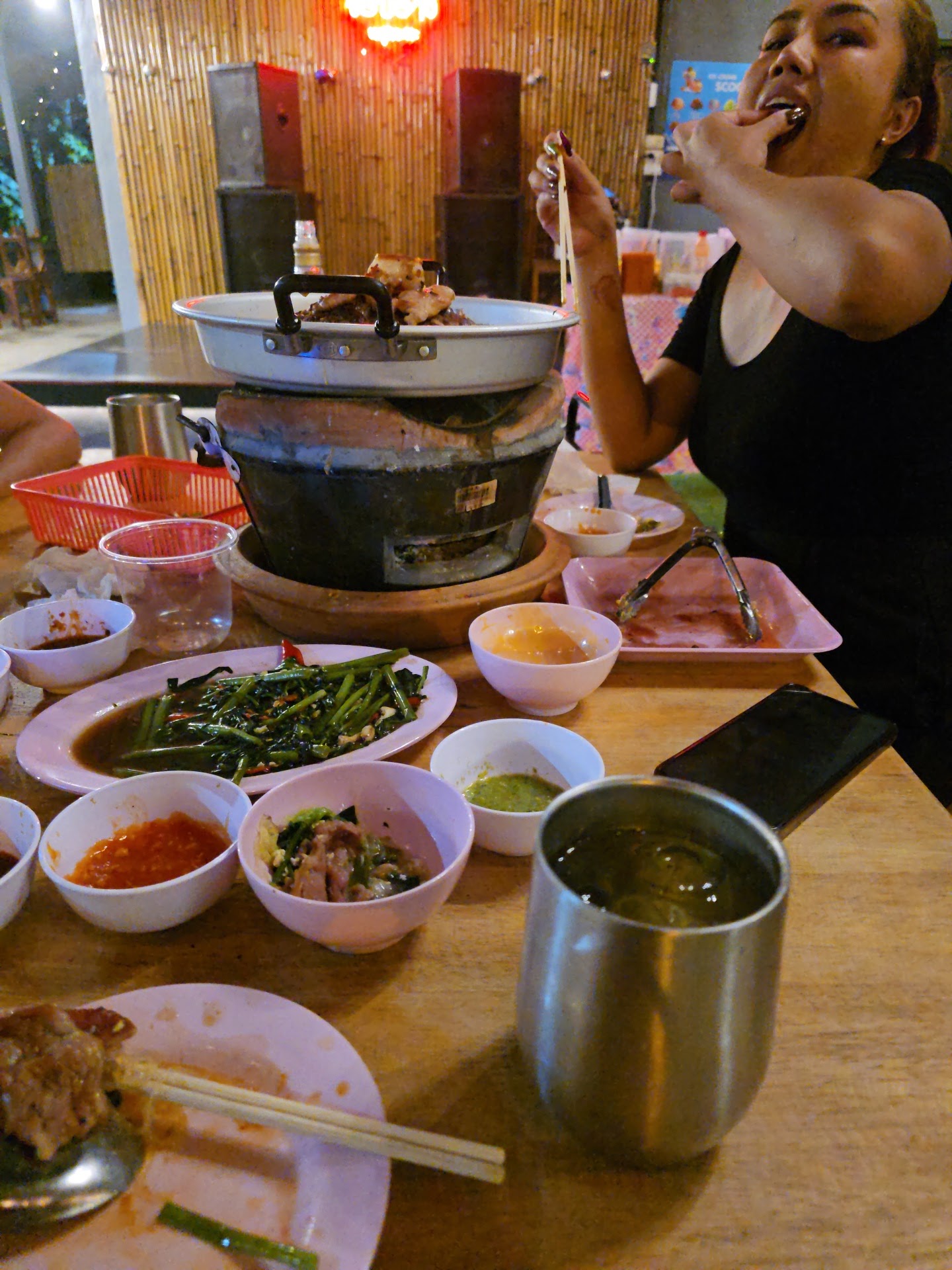 ผู้หญิงกำลังกินหมูกระทะบนโต๊ะอาหารที่เต็มไปด้วยเครื่องเคียง น้ำจิ้ม ผักบุ้งไฟแดง และเตาปิ้งย่างแบบถ่าน