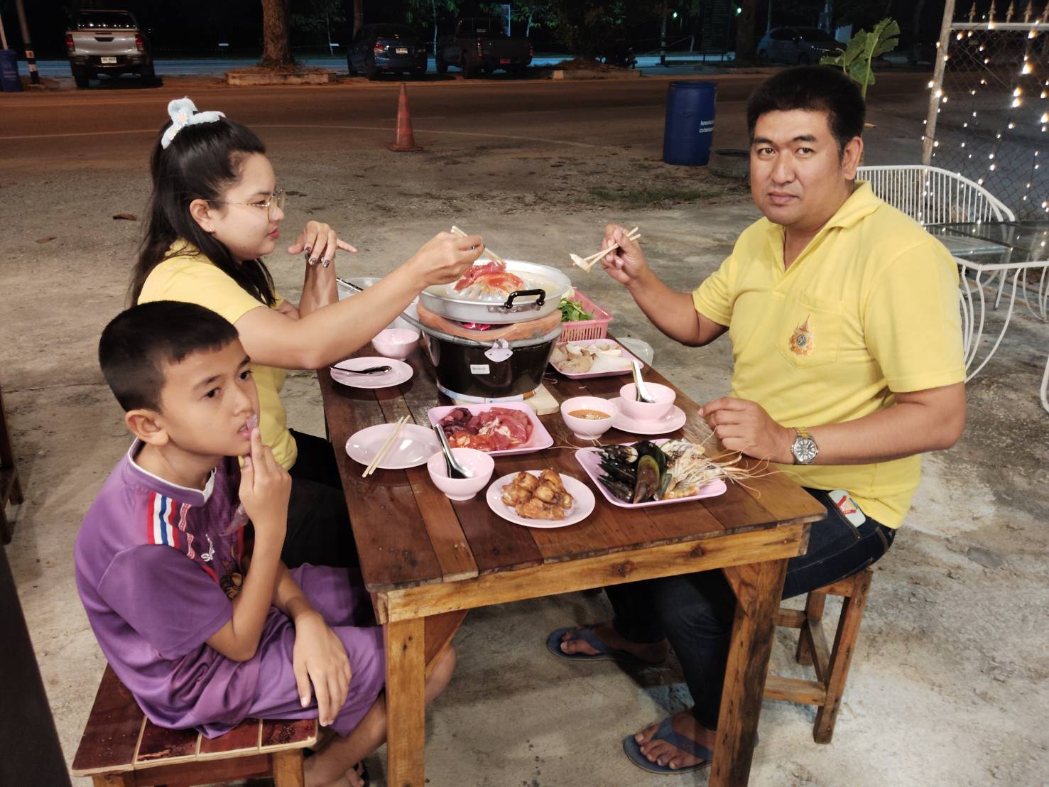 ครอบครัวชาวไทยนั่งทานหมูกระทะกลางแจ้งในตอนกลางคืน มีอาหารทะเลและเนื้อสดหลายจานบนโต๊ะไม้