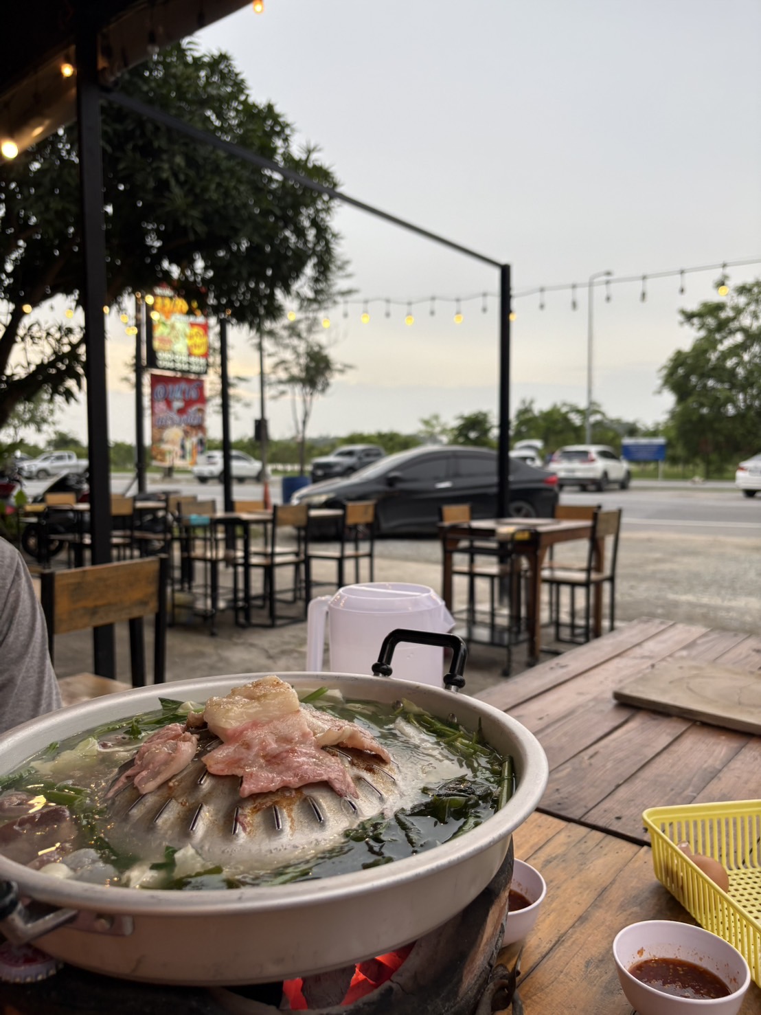 หมูกระทะพร้อมเนื้อหมูและผักบนเตาร้อนที่ร้านอาหารริมถนนบรรยากาศยามเย็นพร้อมไฟประดับ