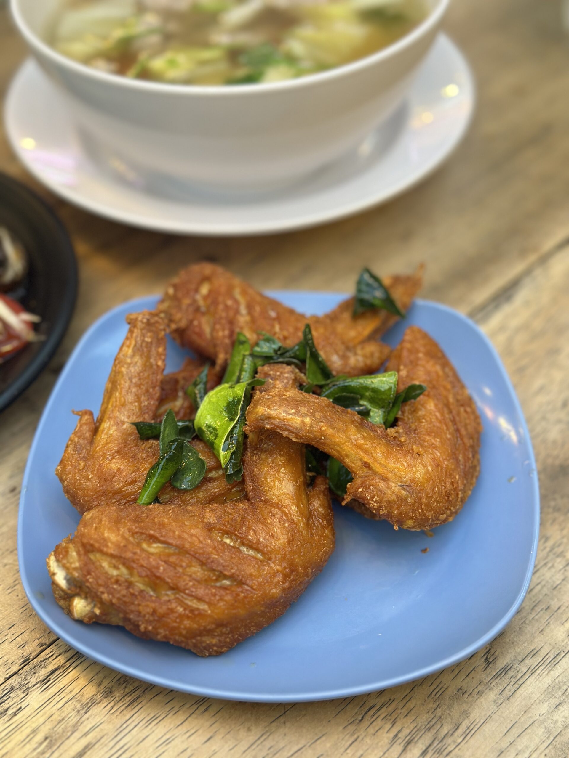 ปีกไก่ทอดกรอบโรยใบโหระพา เสิร์ฟบนจานสีฟ้า พร้อมชามก๋วยเตี๋ยวน้ำใสด้านหลัง