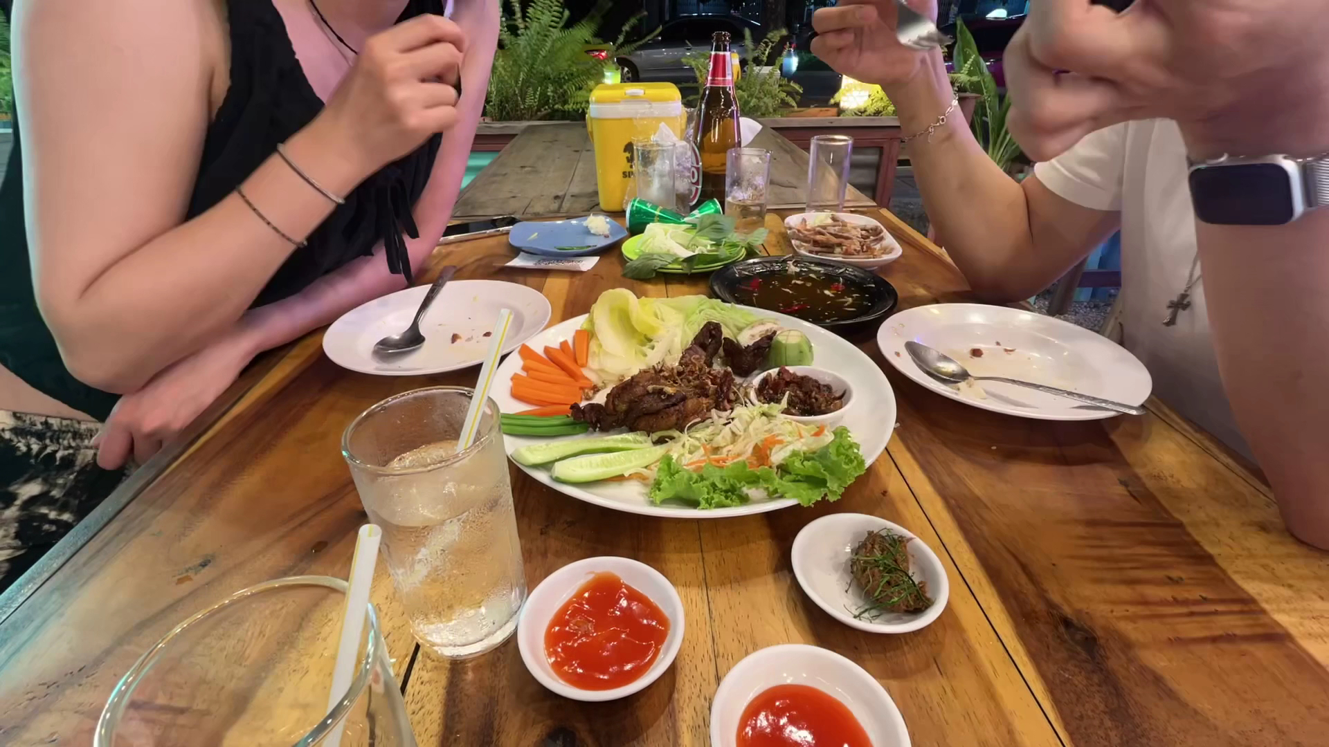 จานเนื้อย่างพร้อมผักสดและน้ำจิ้มบนโต๊ะไม้ในร้านอาหารกลางแจ้ง มีเบียร์และเครื่องดื่มเย็น