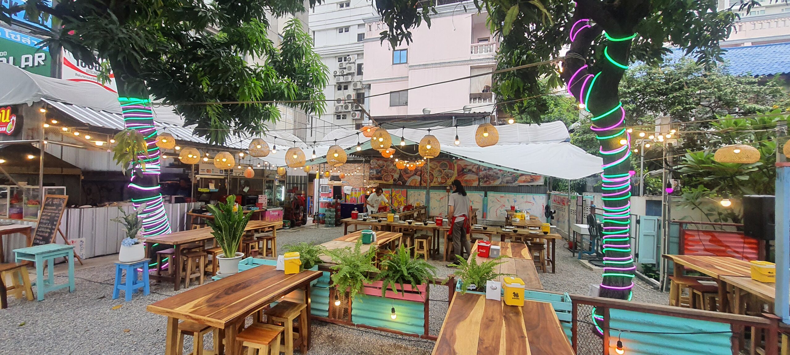 ร้านอาหารกลางแจ้งตกแต่งด้วยไฟนีออนสีเขียวและชมพูพันรอบเสา โคมไฟหวายห้อยระย้า โต๊ะเก้าอี้ไม้เรียงรายใต้ผ้าใบกันแดด บรรยากาศร้านอาหารสไตล์สตรีทฟู้ด