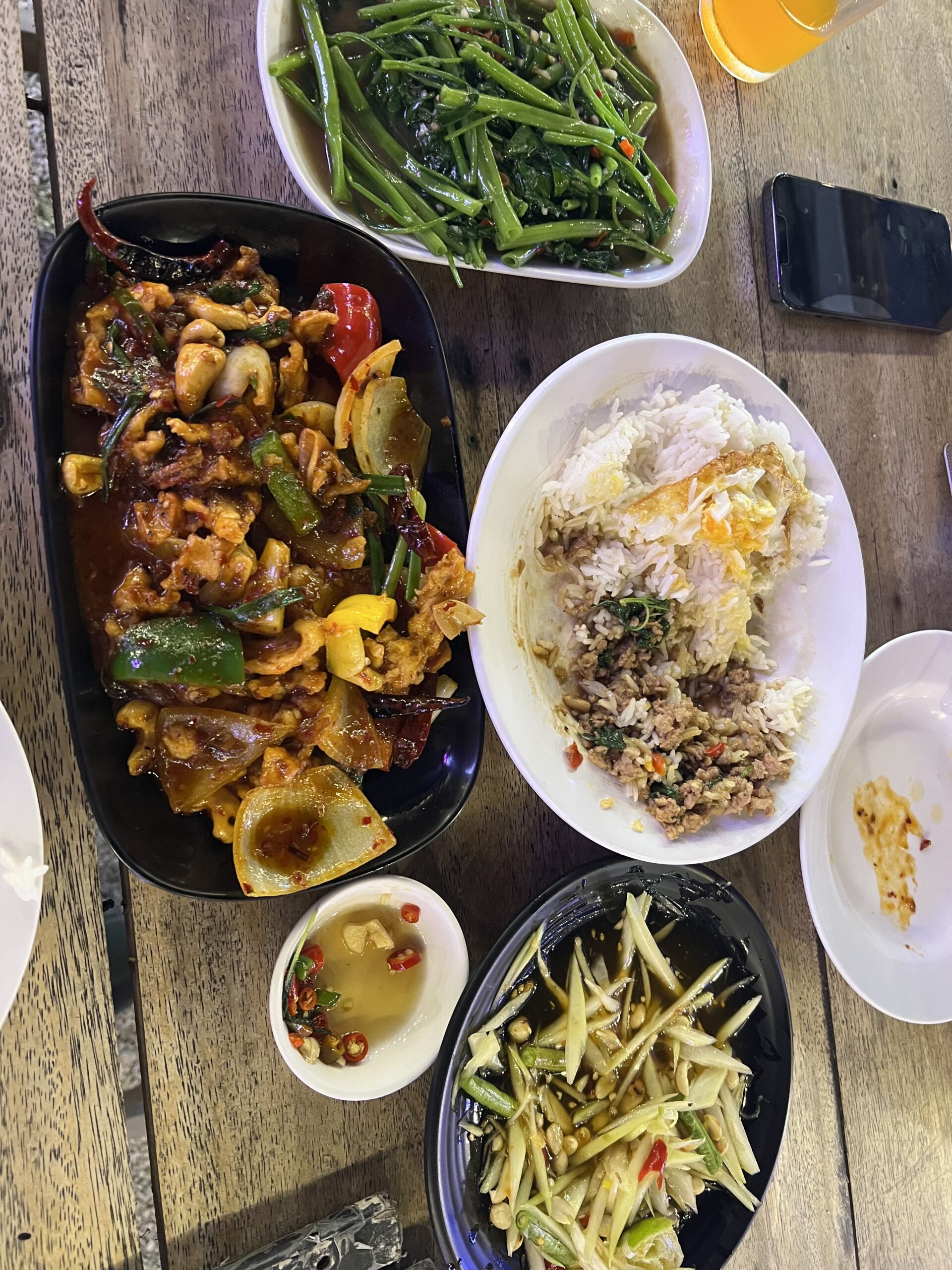 โต๊ะอาหารไทยหลากหลายเมนู มีผัดเผ็ดในจานดำ ข้าวราดกะเพราไข่ดาว ผักบุ้งไฟแดง ส้มตำมะม่วง และน้ำปลาพริก บนโต๊ะไม้