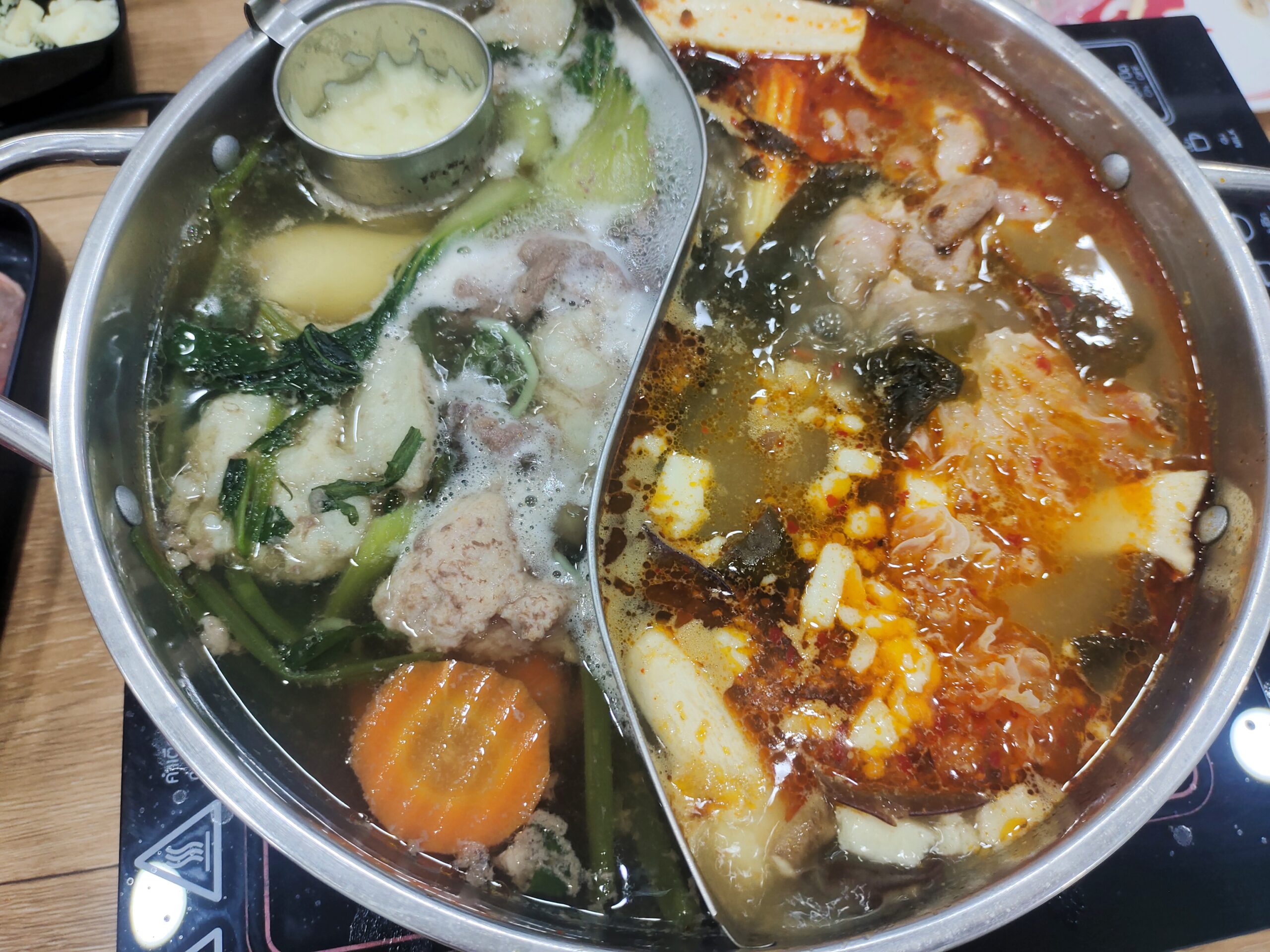 หม้อชาบูสองช่องแบ่งน้ำซุปใสและน้ำซุปเผ็ดแดง มีผักบุ้ง แครอท ลูกชิ้น เต้าหู้ และเนื้อสัตว์ต่างๆ กำลังเดือดบนเตาไฟฟ้า