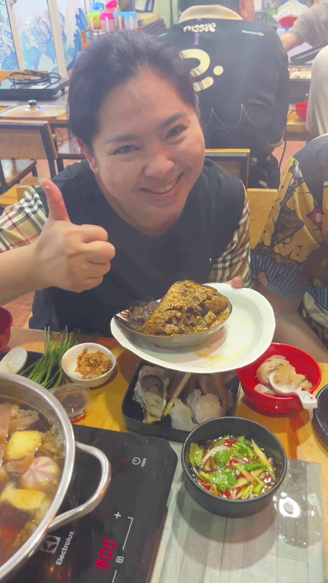 ผู้หญิงยิ้มชูนิ้วโป้งพร้อมจานปูไข่และหม้อไฟสุกี้บนเตาไฟฟ้า Electrolux ในร้านอาหารทะเล