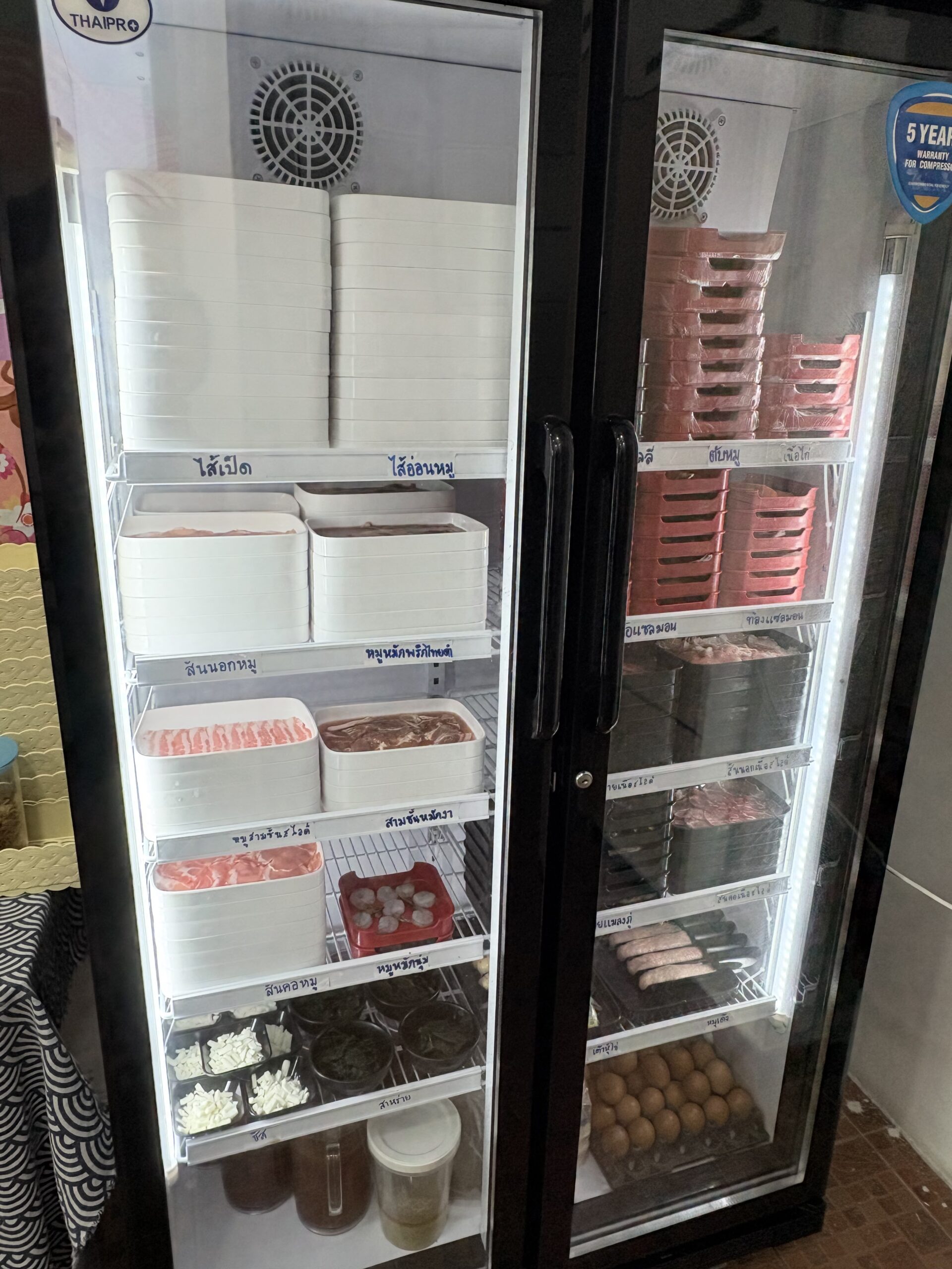 ตู้แช่เย็น THAIPRO แสดงวัตถุดิบปิ้งย่างหลากชนิด เช่น ไส้เป็ด ไส้อ่อนหมู สันนอกหมู หมูหมักพริกไทยดำ สามชั้นหมักยา ไข่ไก่ และไส้กรอก จัดเรียงเป็นระเบียบในถาดโฟม