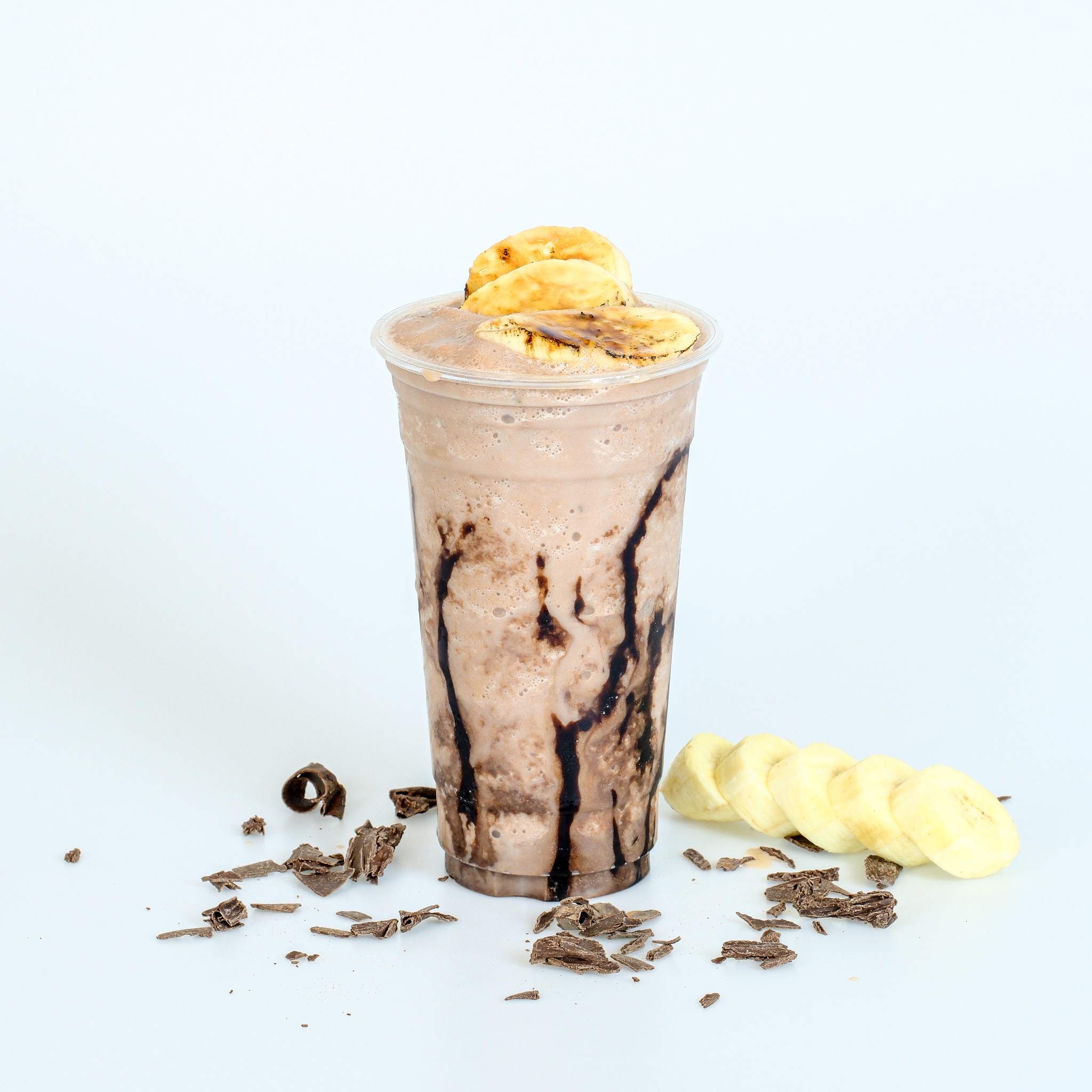 มิลค์เชคช็อกโกแลตกล้วยหอมในแก้วพลาสติกใส ตกแต่งด้วยกล้วยหอมหั่นแว่นและช็อกโกแลตขูดบนพื้นหลังสีขาว