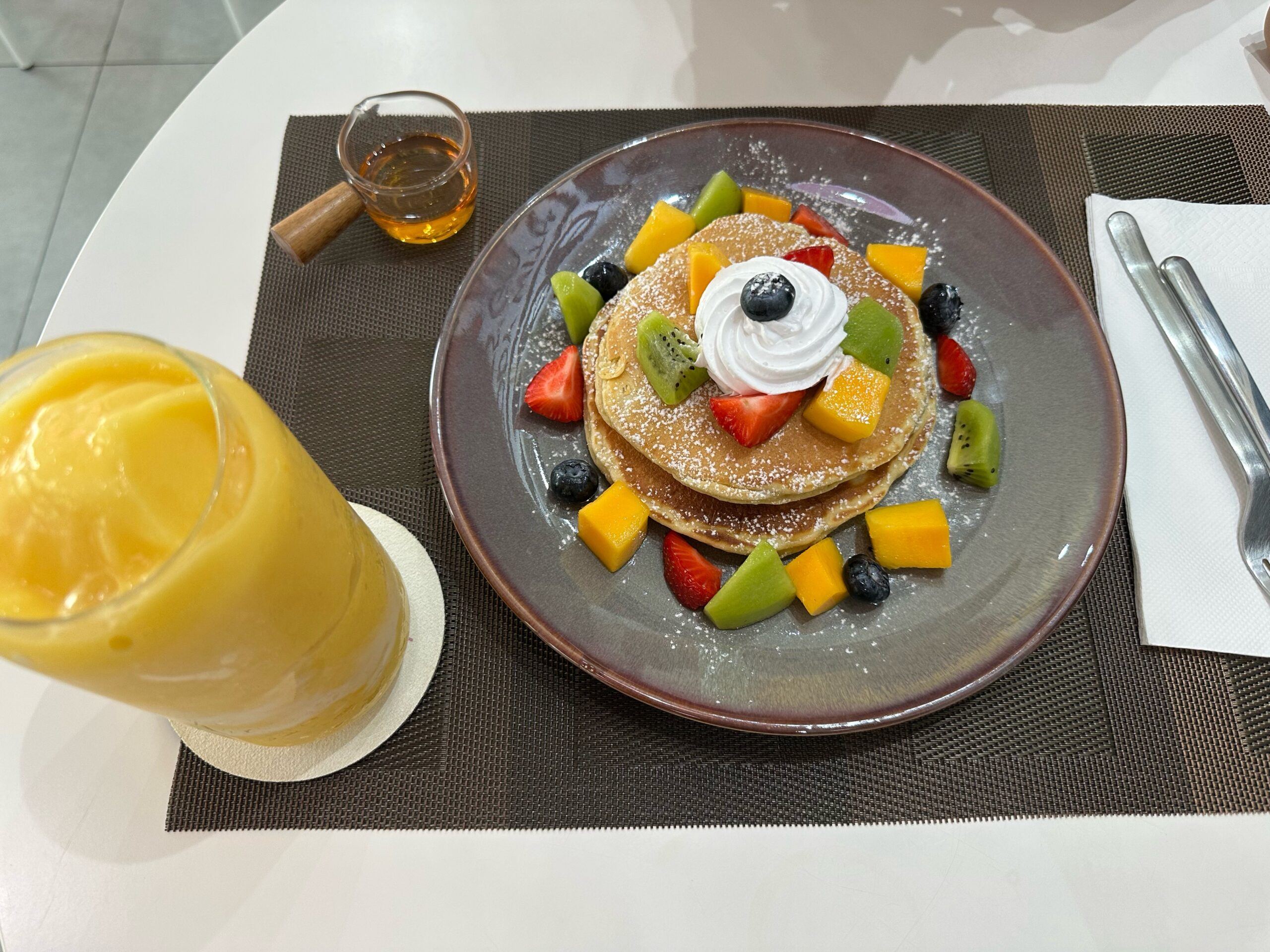 แพนเค้กซ้อนราดน้ำผึ้งพร้อมวิปครีม ผลไม้สด สตรอว์เบอร์รี่ กีวี มะม่วง บลูเบอร์รี่ และสมูทตี้มะม่วงเสิร์ฟบนจานสีเทา