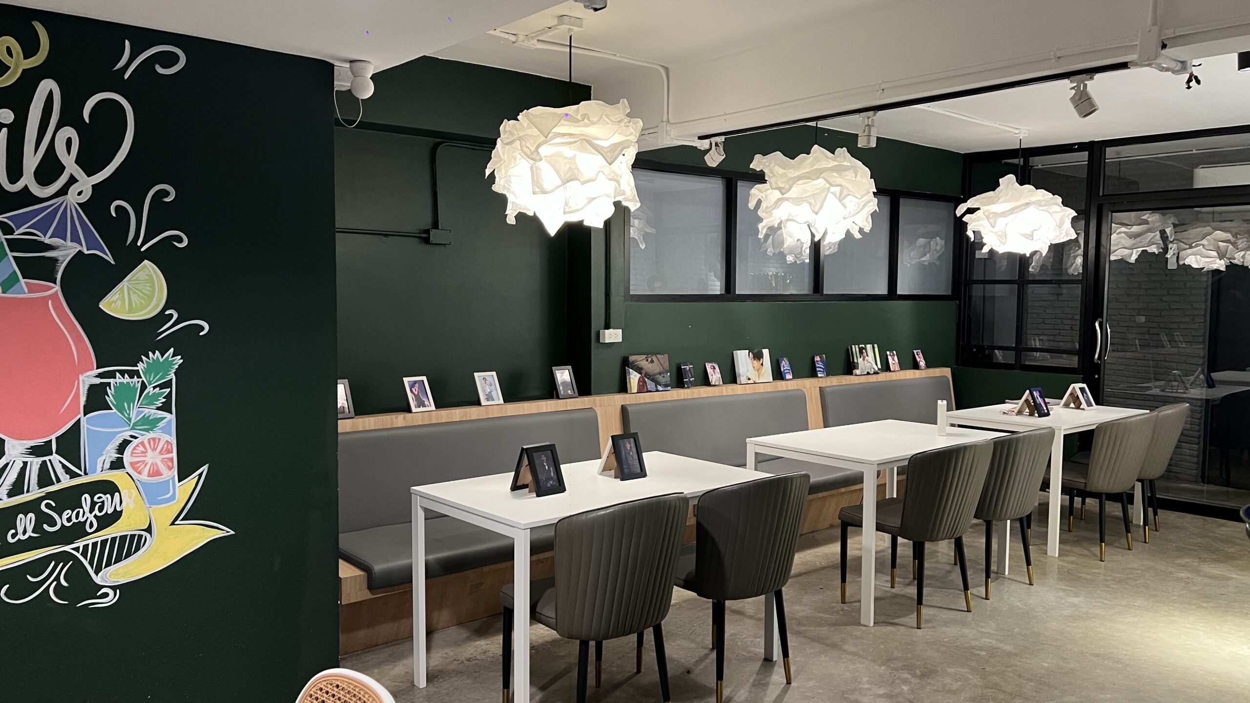 ภายในร้านคาเฟ่สไตล์โมเดิร์นผนังสีเขียวเข้มพร้อมโคมไฟแขวนสีขาวรูปเมฆ โต๊ะสีขาวเก้าอี้สีเทาและภาพถ่ายตกแต่งตามผนัง