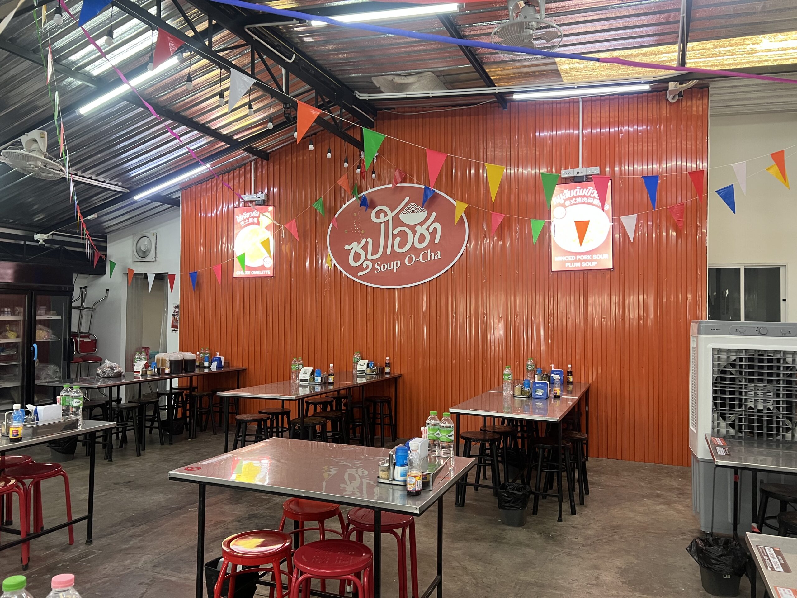 บรรยากาศภายในร้านซุปโอชา มีโต๊ะเก้าอี้หลายชุด ผนังสังกะสีสีส้มพร้อมป้ายโลโก้ร้าน ตกแต่งด้วยธงสามเหลี่ยมสีสันสดใส