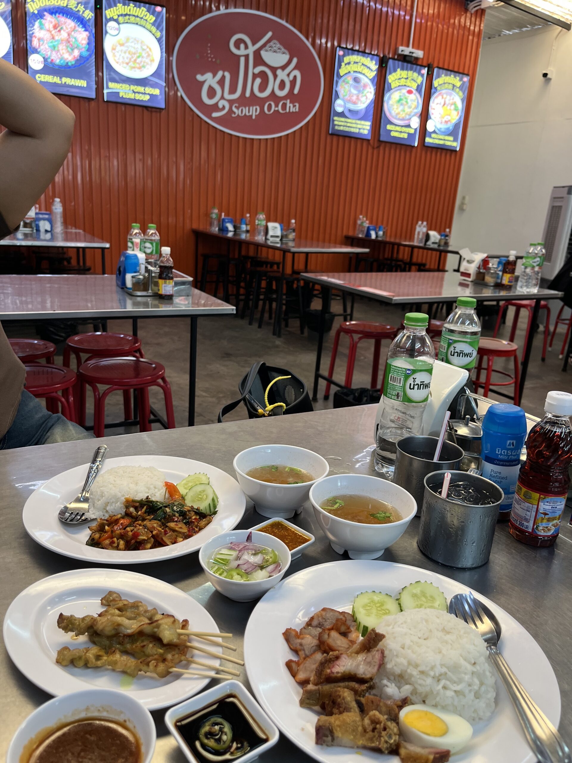 อาหารไทยหลากหลายจานที่ร้านซุปโอชา มีข้าวราดกะเพรา ข้าวหมูแดง หมูสะเต๊ะ และน้ำซุป บนโต๊ะสแตนเลสพร้อมเครื่องดื่ม
