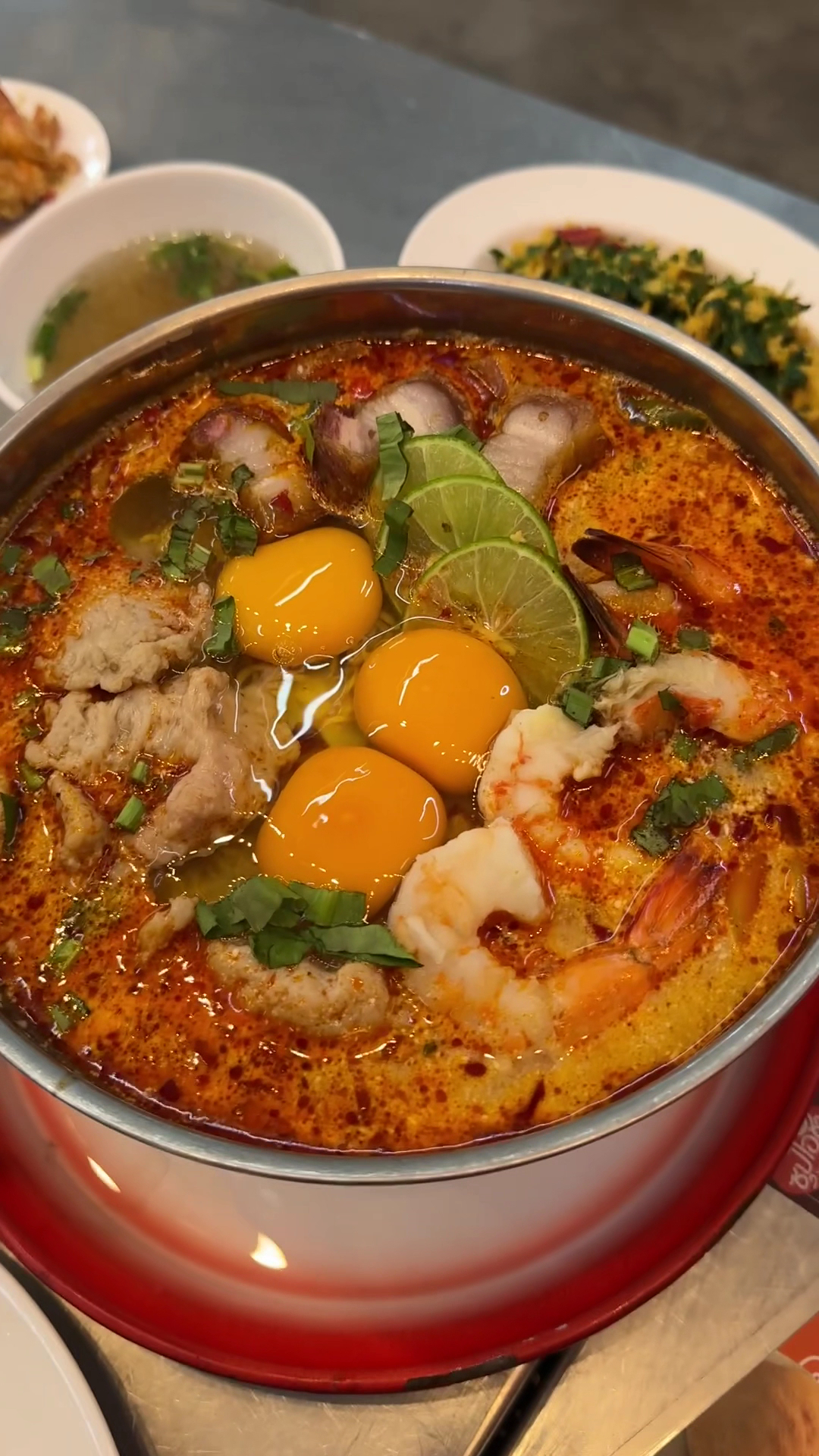 หม้อต้มยำกุ้งน้ำข้นใส่ไข่แดงดิบ มะนาว และเนื้อสัตว์หลายชนิด เสิร์ฟในหม้อสแตนเลสบนถาดแดง