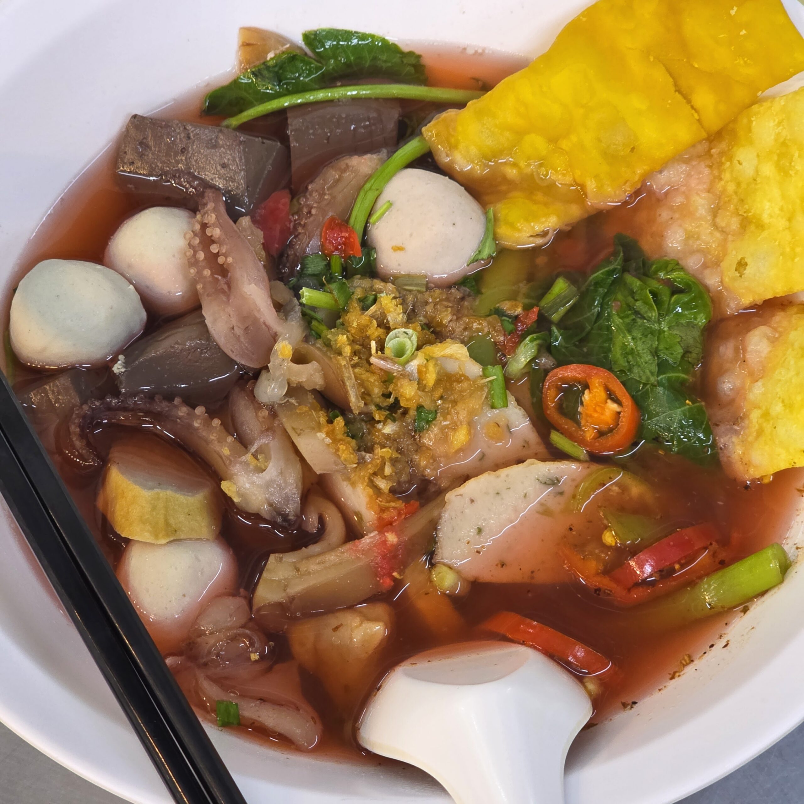 ชามเย็นตาโฟน้ำแดงใส่ลูกชิ้นปลา เต้าหู้ทอด ปลาหมึก เลือดหมู ผักบุ้ง และพริกสด