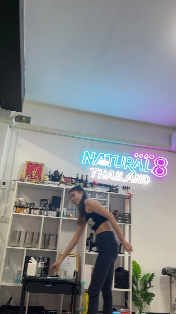 พนักงานสาวในชุดดำกำลังทำงานภายในร้าน Natural8 Thailand ที่มีป้ายไฟนีออนสีฟ้าและชมพูเป็นสัญลักษณ์