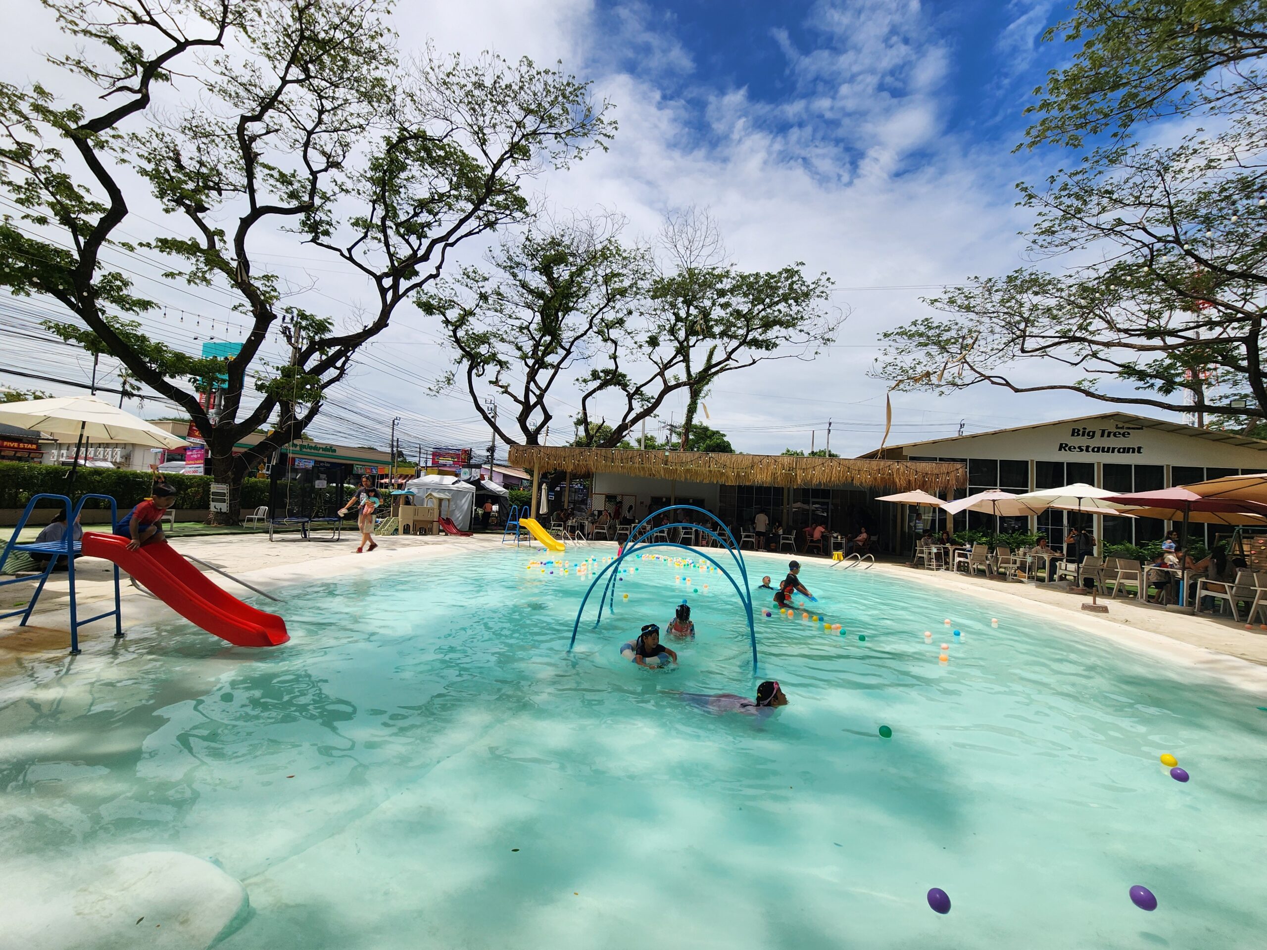 ร้าน Big Tree Restaurant พร้อมสระว่ายน้ำสำหรับเด็กพร้อมสไลเดอร์สีแดงและน้ำพุ ล้อมรอบด้วยต้นไม้ใหญ่และร่มกันแดด