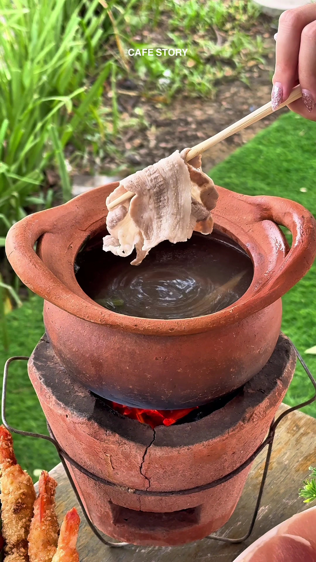 หม้อดินเผาตั้งบนเตาถ่านแดง มีชิ้นเนื้อหมูสไลด์คีบด้วยตะเกียบพร้อมลวกในน้ำซุปร้อน สไตล์ชาบูหม้อดิน