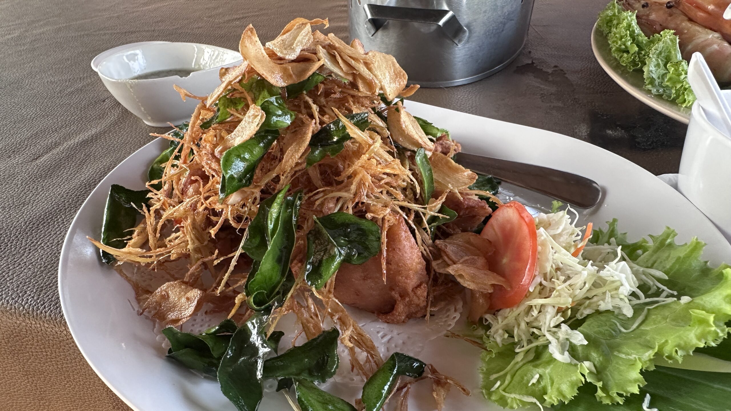 ปลาทอดกรอบโรยสมุนไพรใบกะเพราและกระเทียมเจียวบนจานขาวพร้อมผักสลัดและมะเขือเทศ