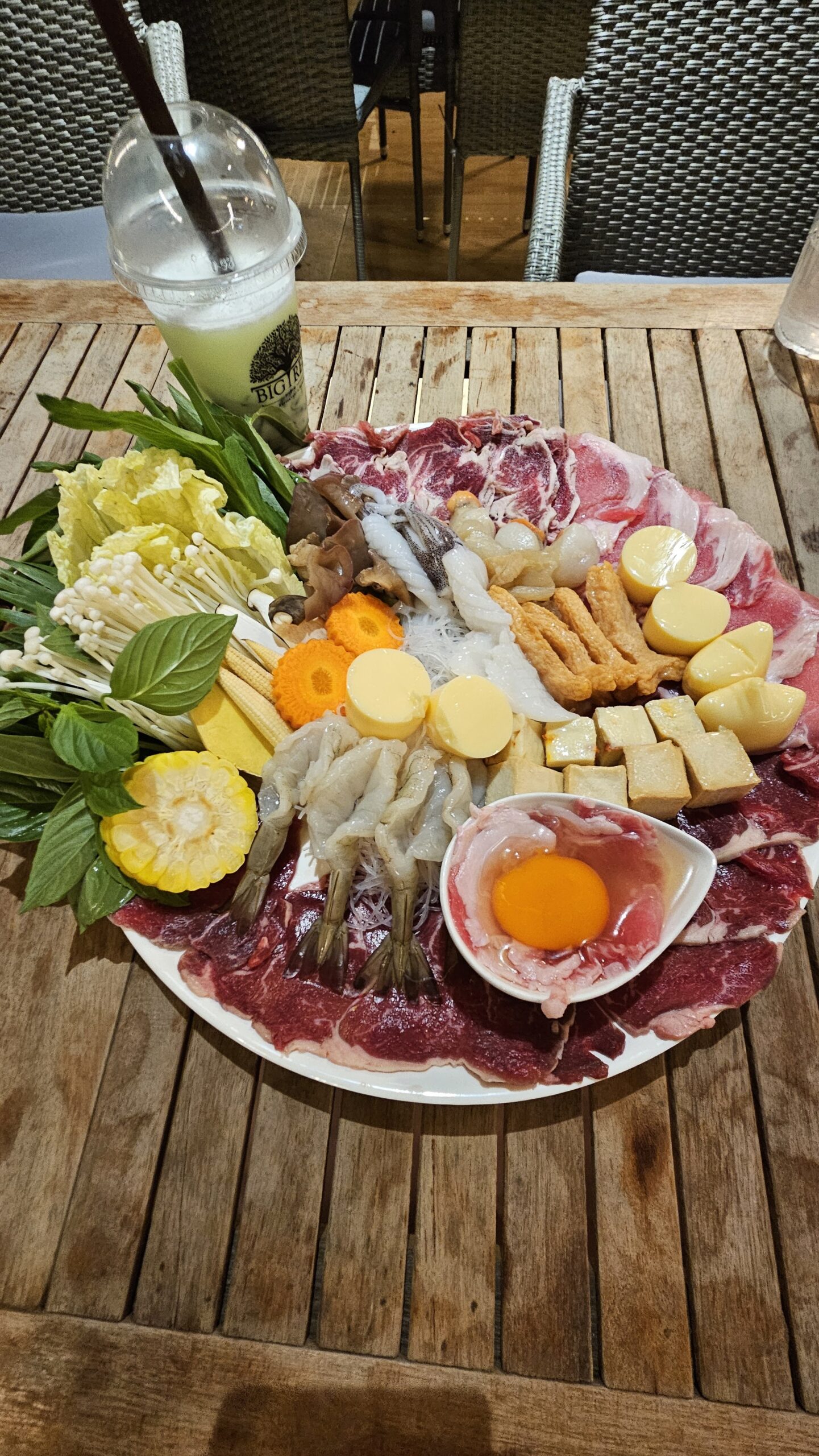 จานวัตถุดิบสำหรับชาบูหรือสุกี้ มีเนื้อวัวสไลด์ กุ้งสด เห็ดเข็มทอง ข้าวโพดอ่อน ผักบุ้ง เต้าหู้ ไข่แดง และผักสดต่างๆ บนโต๊ะไม้ในร้านอาหาร