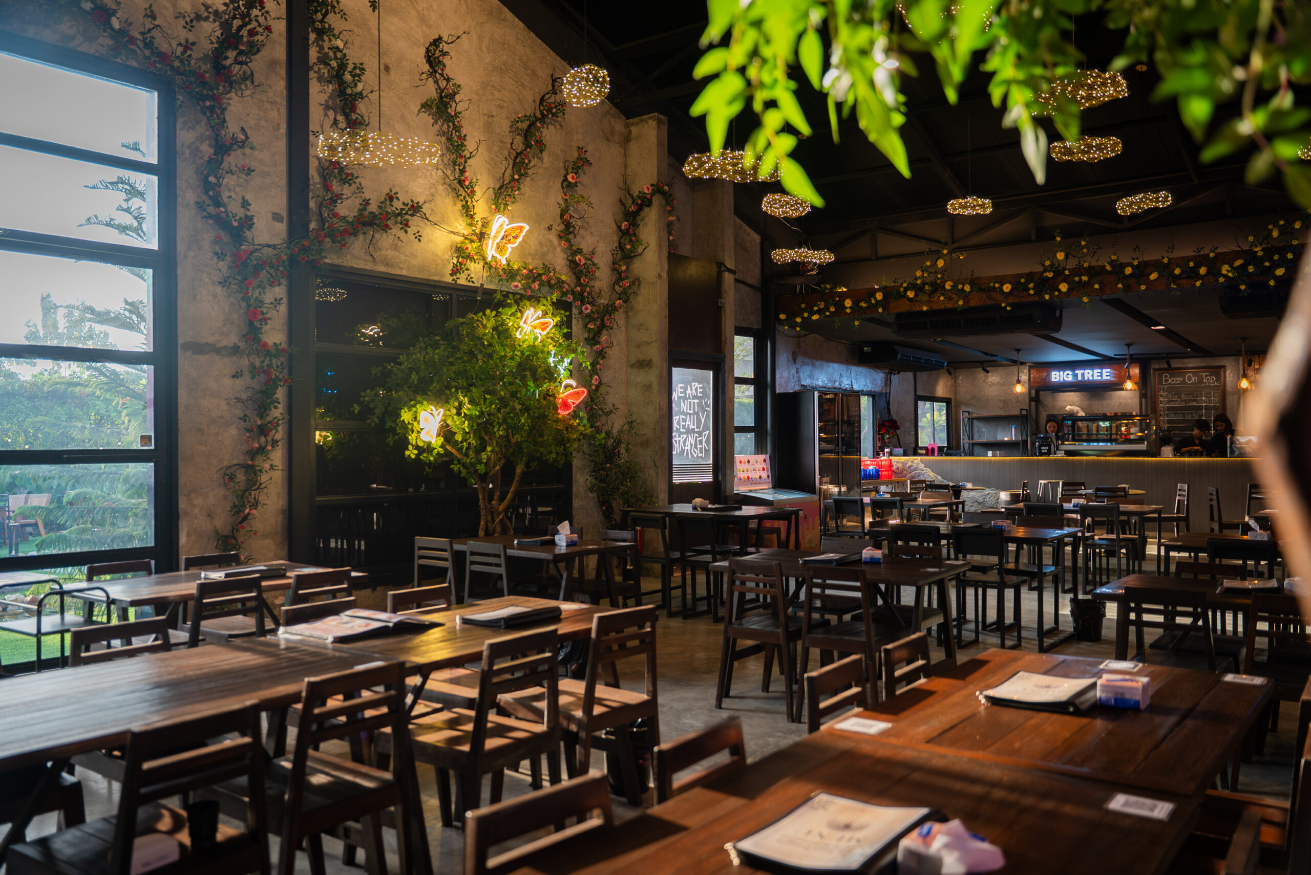 บรรยากาศภายในร้าน Big Tree ตกแต่งด้วยต้นไม้ ดอกไม้ประดับ ผีเสื้อนีออน และไฟตกแต่งเพดาน พร้อมโต๊ะไม้จัดเรียงอย่างสวยงาม