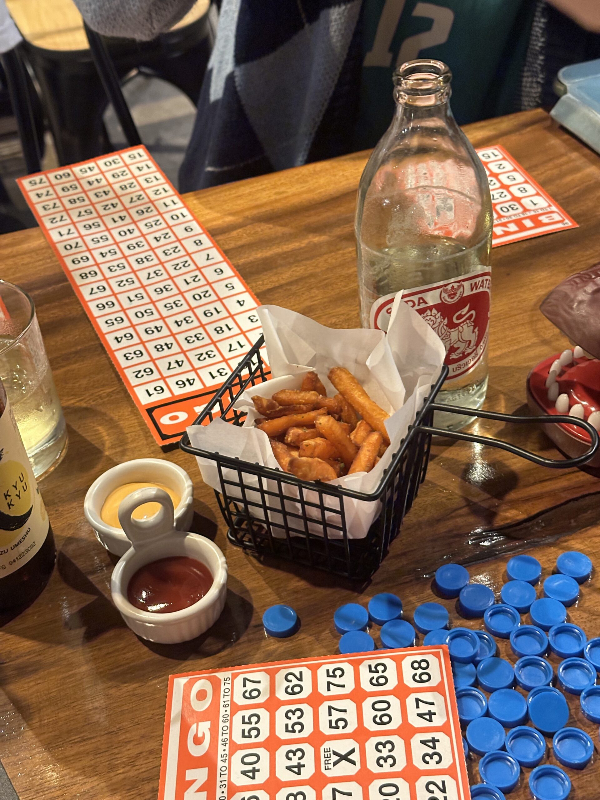 โต๊ะอาหารในร้านอาหารที่มีเฟรนช์ฟรายส์ในตะกร้าเหล็ก ซอสมะเขือเทศและซอสชีส ขวดโซดา บัตรบิงโกสีแดง และชิปสีน้ำเงินสำหรับเล่นบิงโก