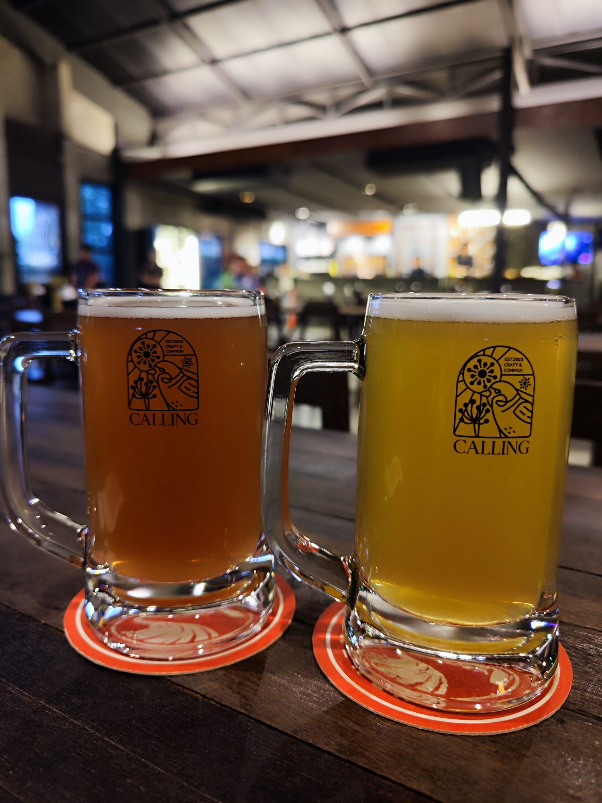 แก้วเบียร์คราฟท์สองใบยี่ห้อ Calling Craft & Common วางบนโต๊ะไม้พร้อมที่รองแก้วสีแดง ในบรรยากาศร้านเบียร์