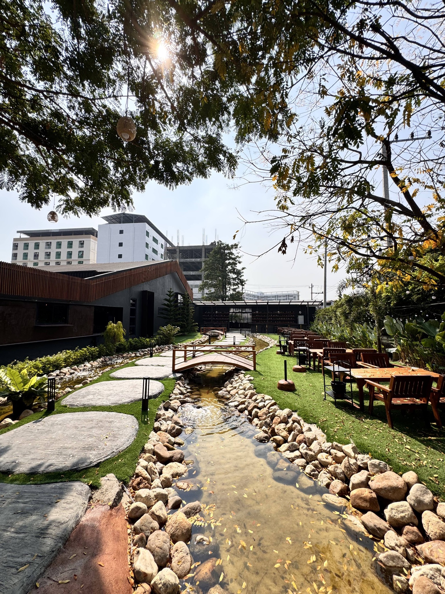 บรรยากาศร้านอาหารกลางแจ้งพร้อมลำธารหินธรรมชาติ สะพานไม้ สนามหญ้าเขียวขจี และโต๊ะเก้าอี้ไม้เรียงรายท่ามกลางต้นไม้ใหญ่ยามแสงแดดอ่อน