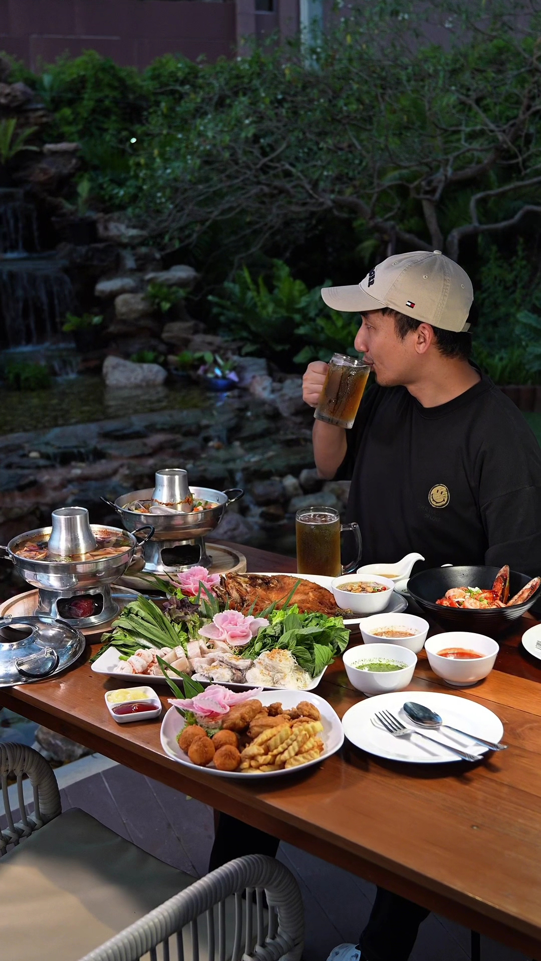 ชายสวมหมวกนั่งดื่มเบียร์ที่โต๊ะอาหารริมน้ำตกจำลอง เต็มไปด้วยอาหารทะเลหม้อไฟ ปลาทอด ของทอด และน้ำจิ้มหลากชนิด