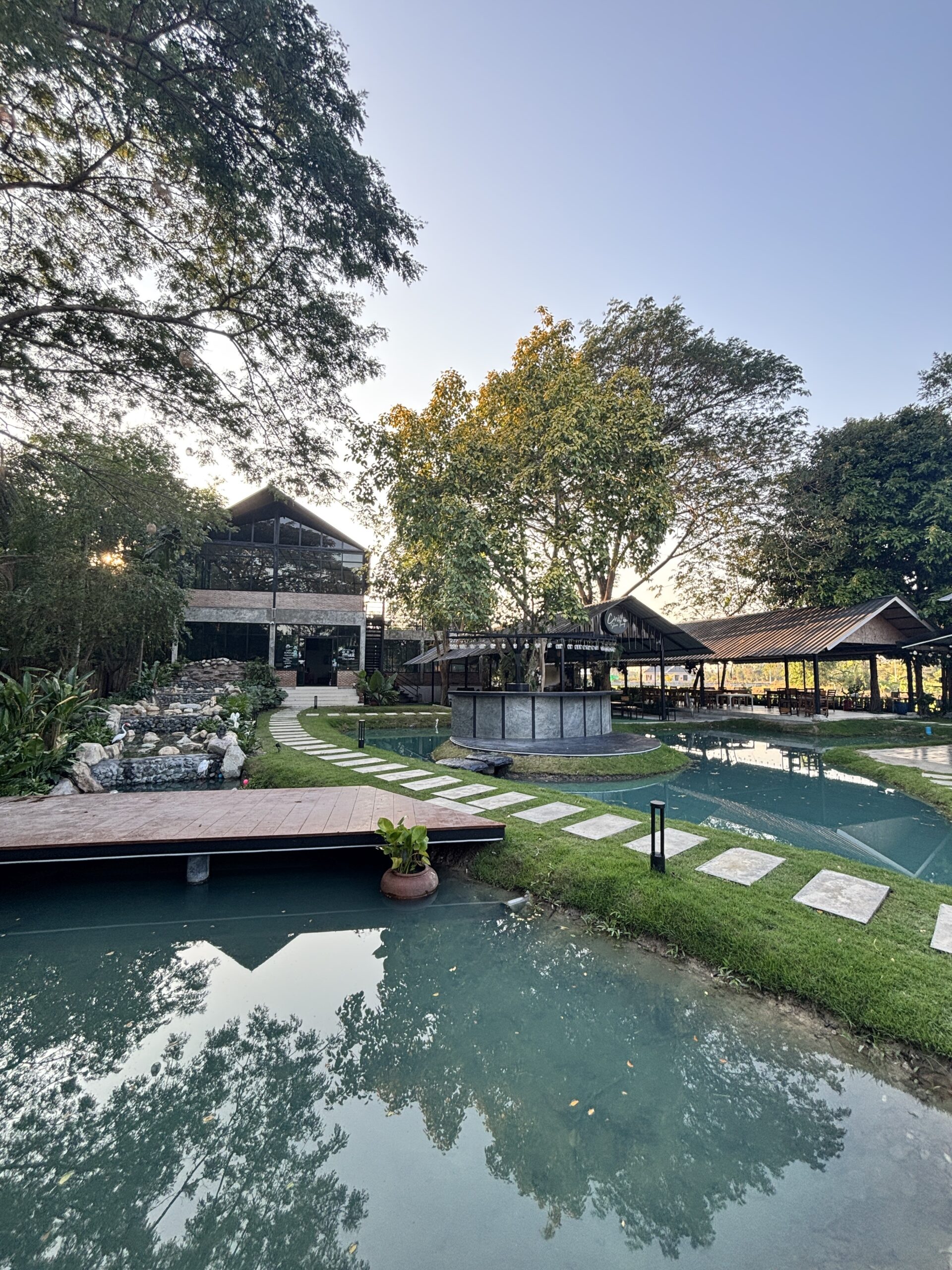 ร้านอาหารริมน้ำพร้อมสระน้ำสีเขียว ศาลาหลังคาจั่ว ทางเดินหินบนสนามหญ้า และเวทีกลางน้ำ ล้อมรอบด้วยต้นไม้ใหญ่ในบรรยากาศยามเย็น