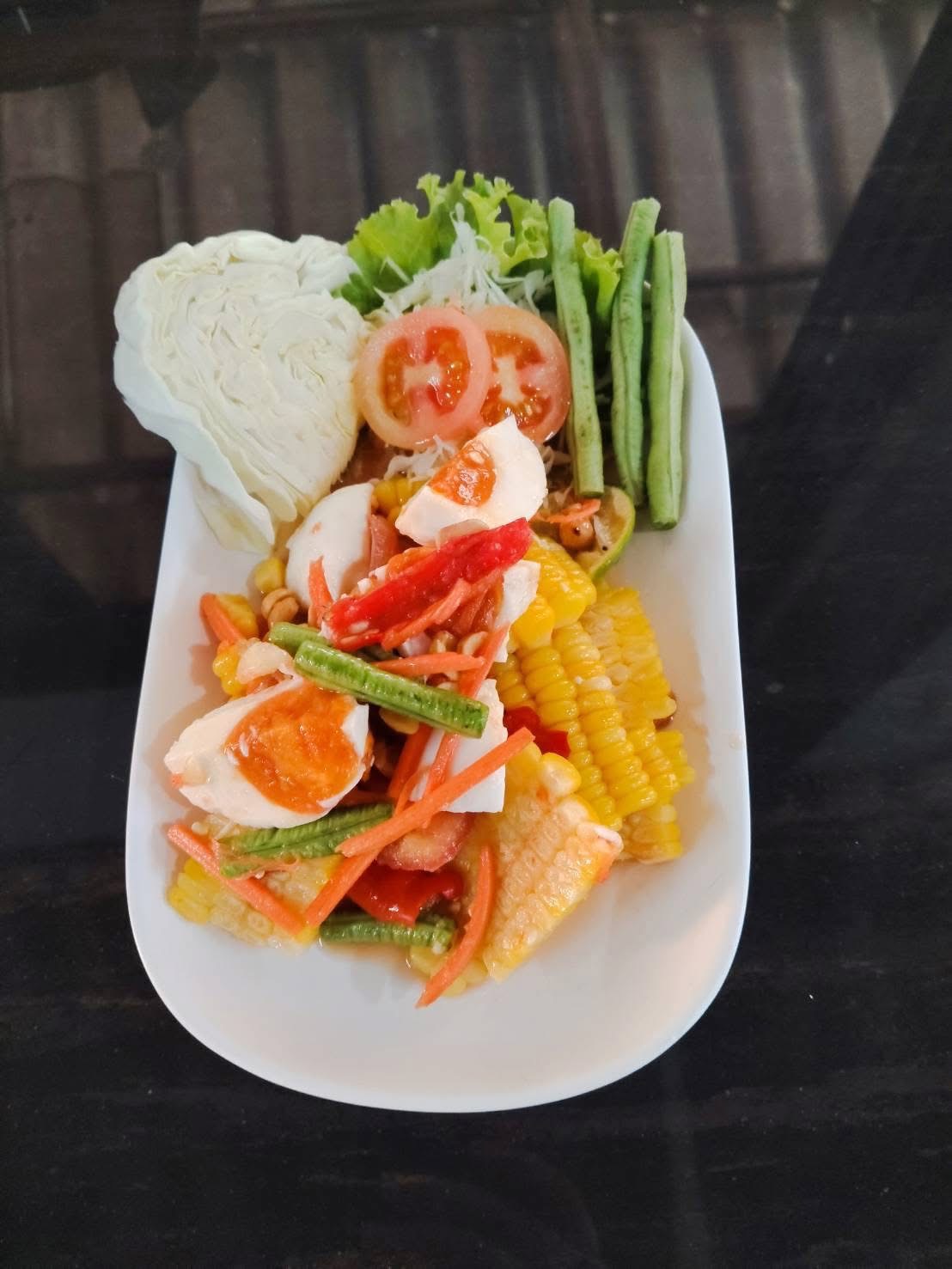 ส้มตำข้าวโพดไข่เค็ม เสิร์ฟพร้อมกะหล่ำปลี ถั่วฝักยาว มะเขือเทศ และผักสด บนจานสีขาว