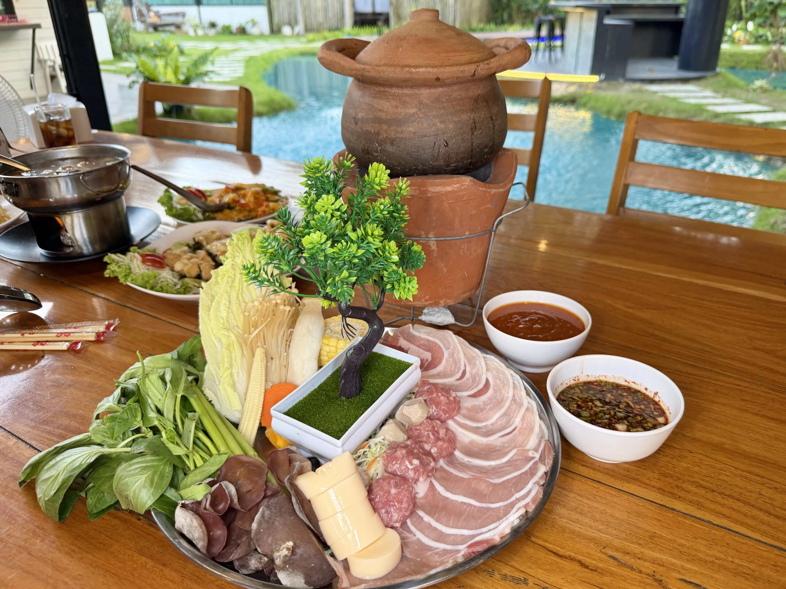จานหมูสไลด์และผักสดพร้อมหม้อดินสำหรับจิ้มจุ่มบนโต๊ะไม้ริมสระว่ายน้ำ พร้อมน้ำจิ้มสองถ้วย