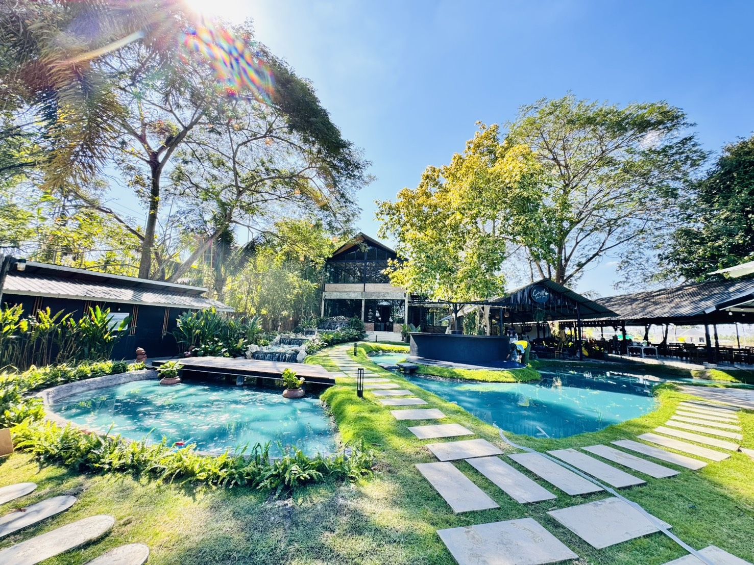 คาเฟ่สวยท่ามกลางสวนเขียวขจีพร้อมสระน้ำสีฟ้าใส ทางเดินหินปูนท่ามกลางสนามหญ้า และอาคารสไตล์โมเดิร์นลอฟท์ในบรรยากาศแดดอุ่น