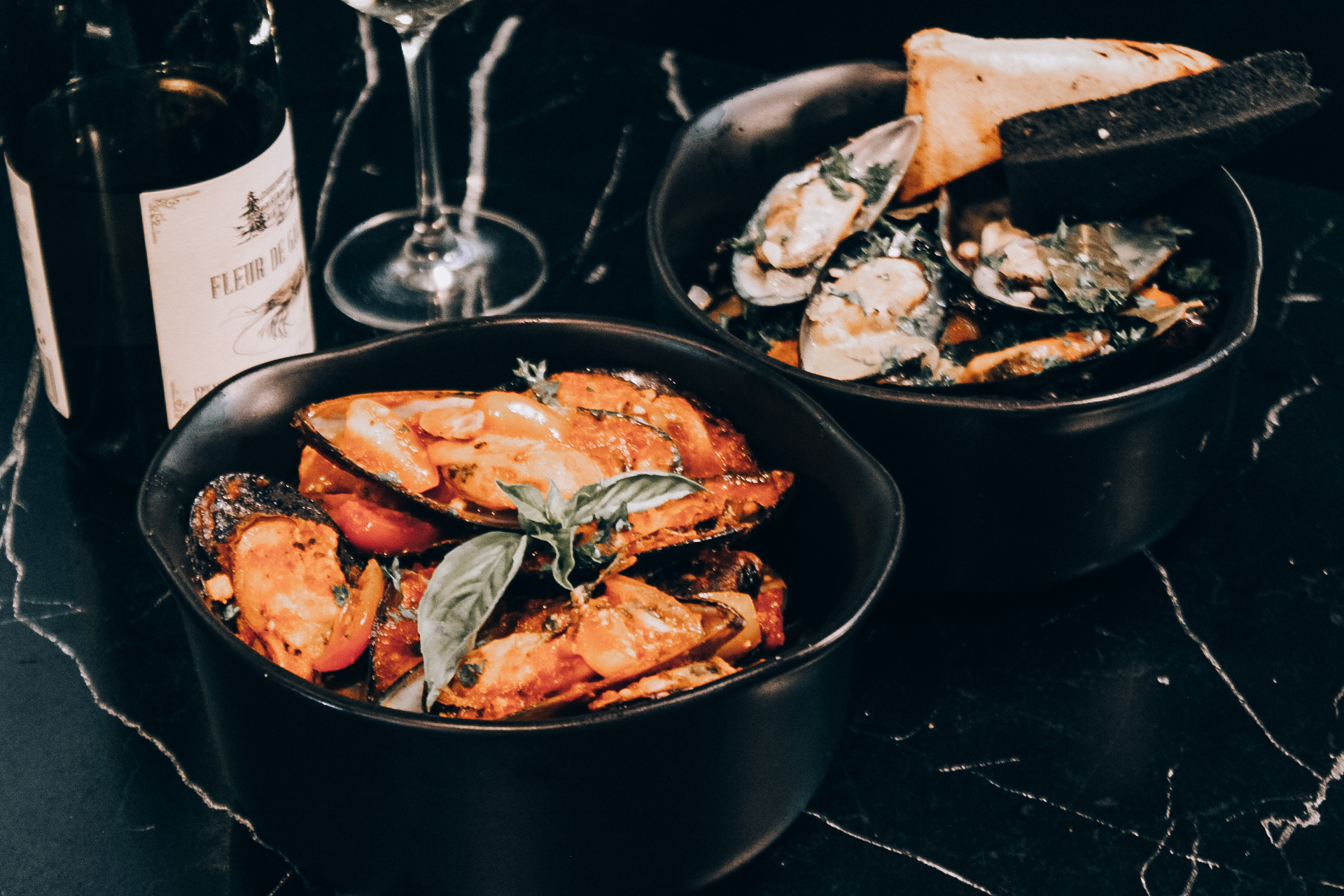 หอยแมลงภู่ในซอสมะเขือเทศพร้อมใบโหระพาเสิร์ฟในชามดำคู่กับขนมปังและไวน์บนโต๊ะหินอ่อนสีดำ