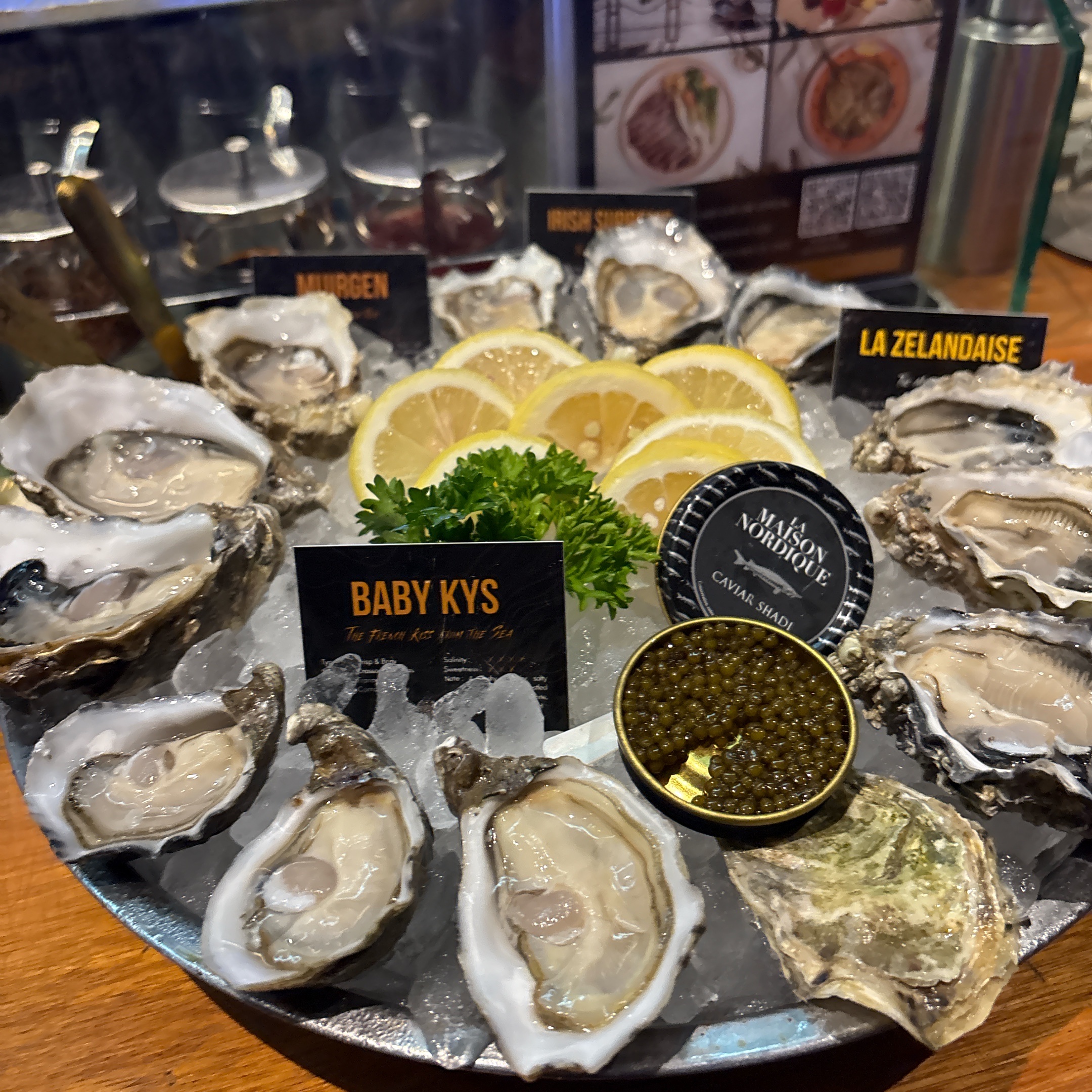จานหอยนางรมสดหลากหลายชนิดบนน้ำแข็ง พร้อมคาเวียร์ La Maison Nordique มะนาวฝานและผักชีฝรั่ง มีป้ายชื่อ Baby Kys, Murgen, Irish และ La Zelandaise