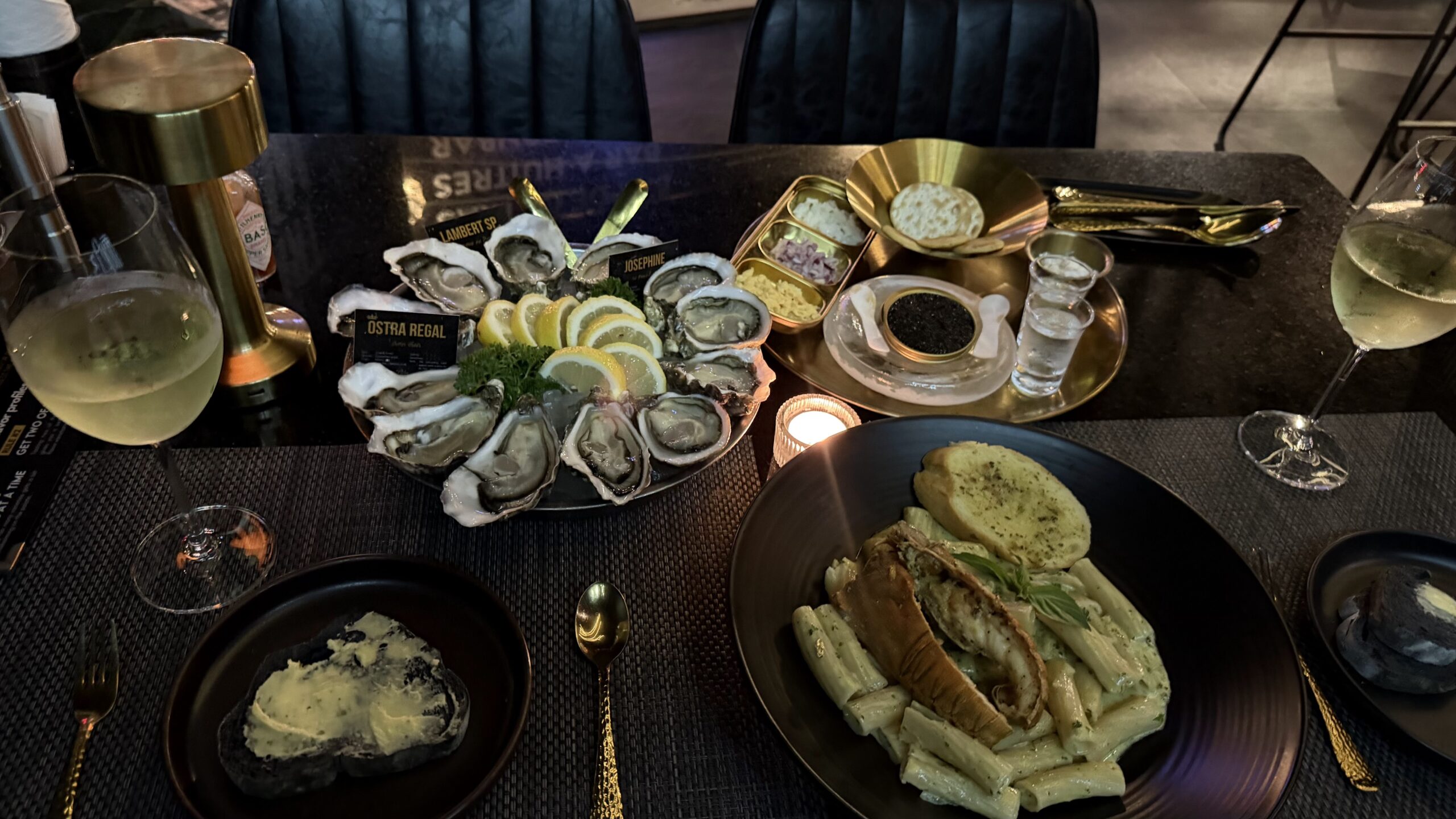 จานหอยนางรมสดเสิร์ฟบนจานใหญ่พร้อมมะนาวฝานบาง คาเวียร์ พาสต้าครีมซอสกุ้ง และไวน์ขาว บนโต๊ะอาหารหรูหราตกแต่งด้วยภาชนะสีทอง