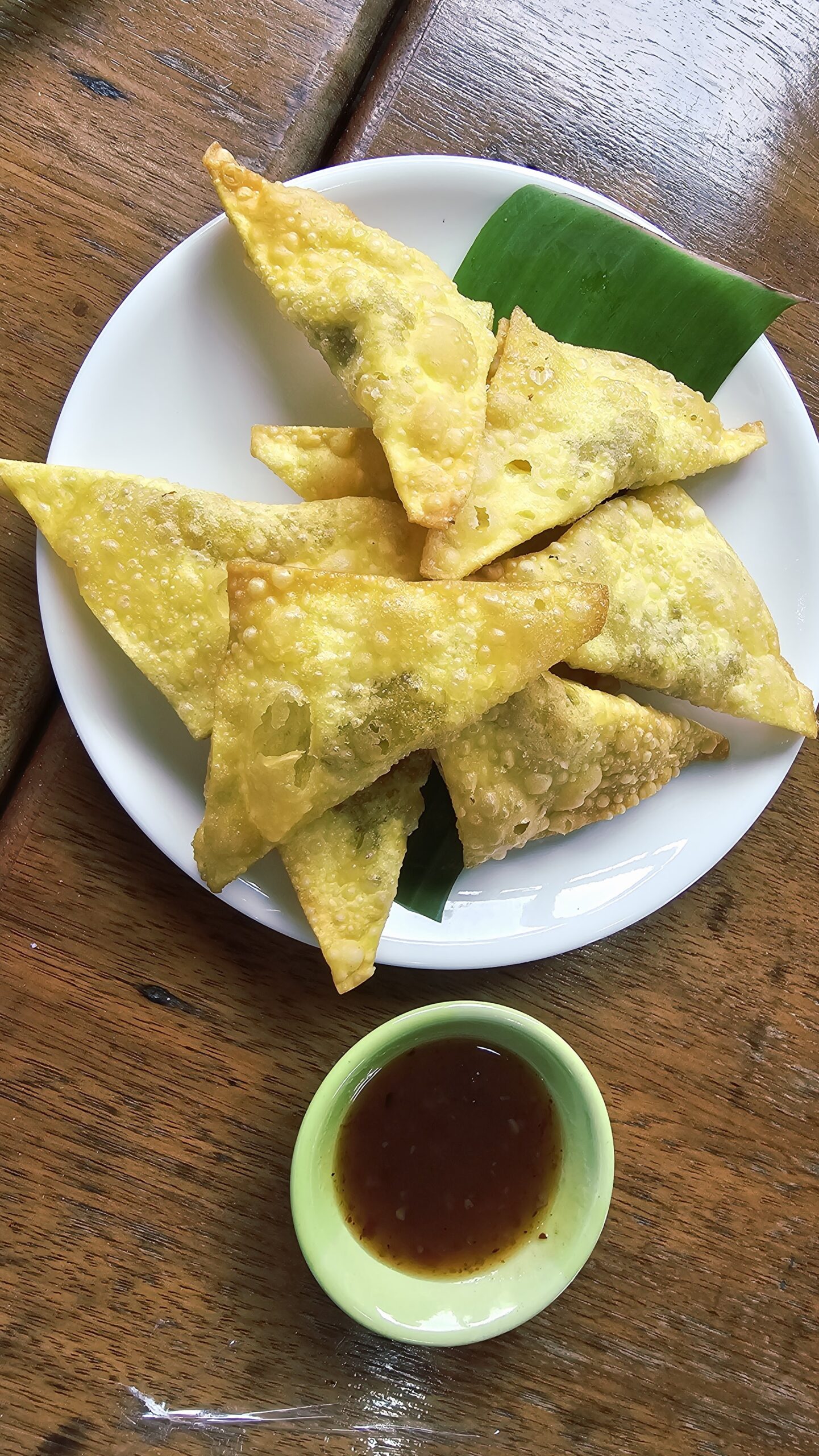 เกี๊ยวทอดกรอบสีทองเสิร์ฟบนจานขาวรองใบตองพร้อมน้ำจิ้มเปรี้ยวหวานในถ้วยเขียว