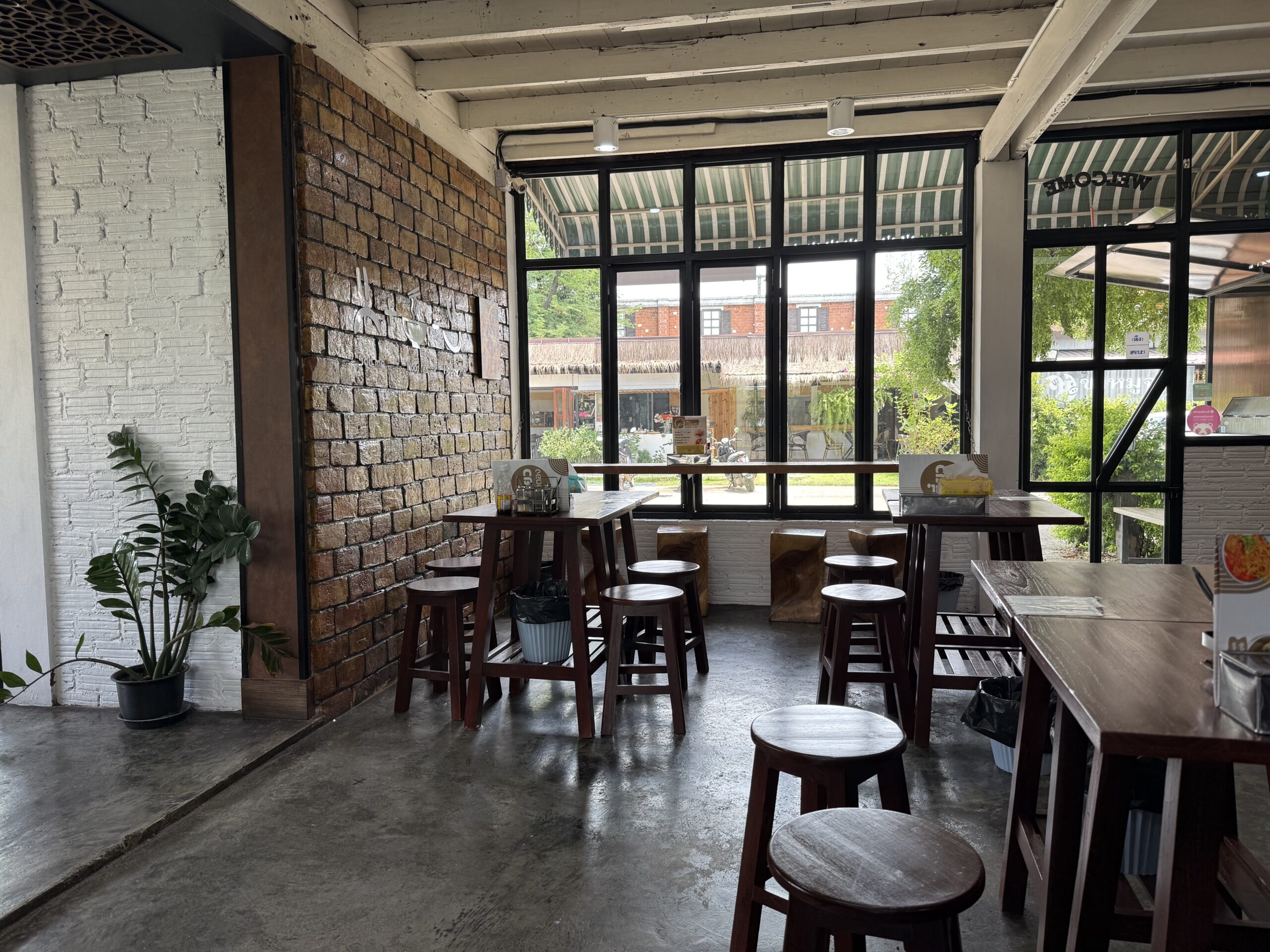 ภายในร้านอาหารสไตล์อินดัสเทรียลลอฟท์ มีผนังอิฐเก่า เก้าอี้สตูลไม้ โต๊ะไม้ หน้าต่างกระจกบานใหญ่ พื้นปูนขัดมัน และต้นไม้ประดับ