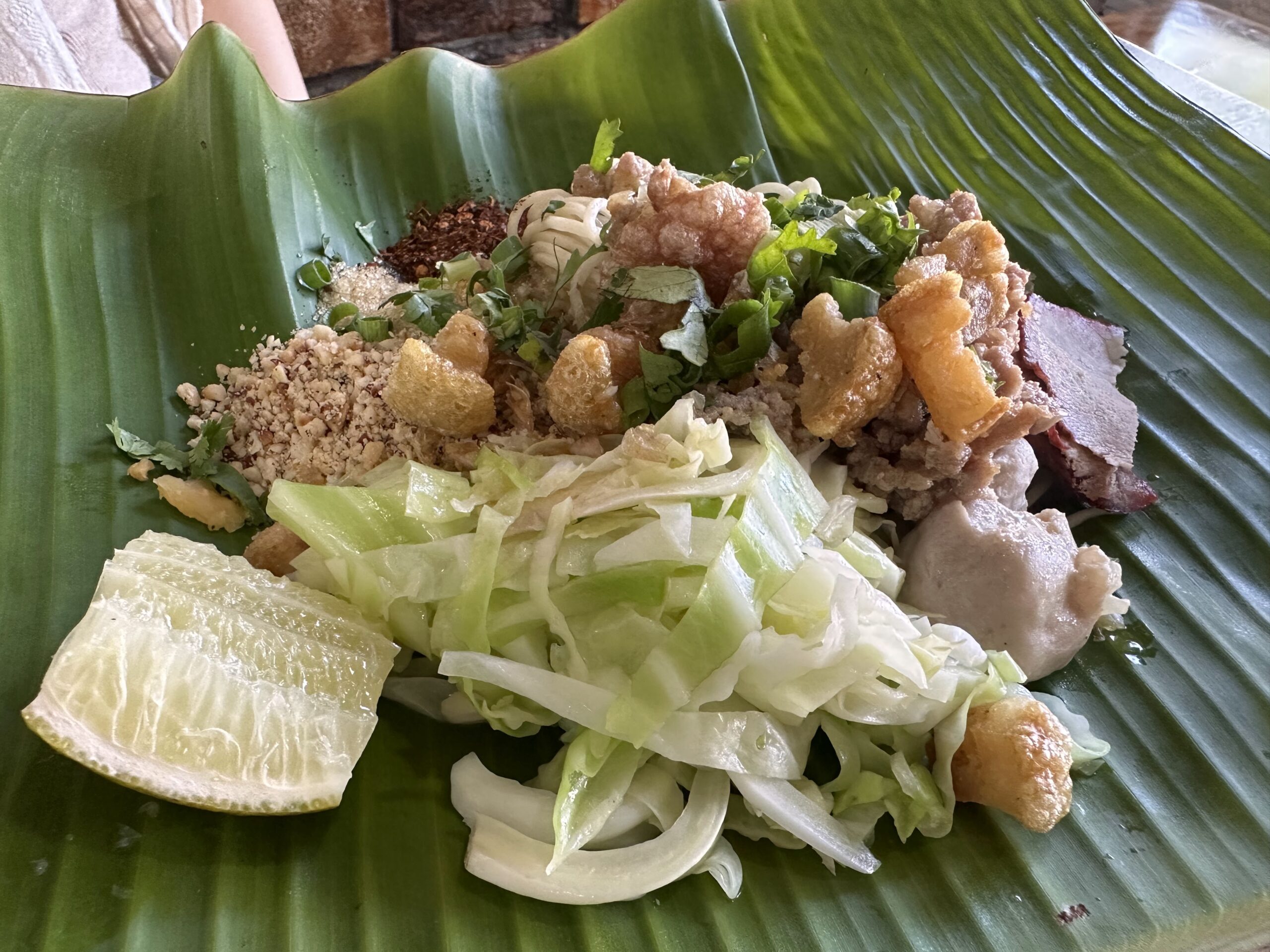 เส้นใหญ่หมูกรอบบนใบตอง พร้อมผักกาดขาว ถั่วลิสงป่น พริกป่น มะนาว และผักชี