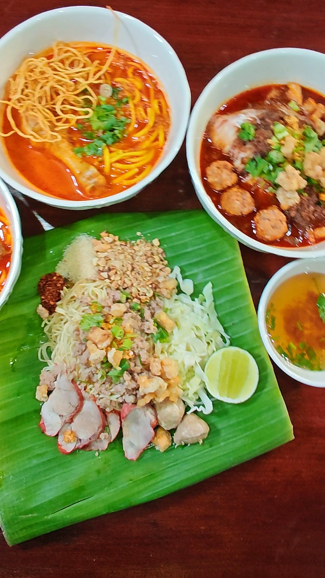 อาหารไทยหลากหลายบนโต๊ะไม้ ข้าวซอย ก๋วยเตี๋ยวน้ำตก บะหมี่แห้งหมูแดงบนใบตอง พร้อมน้ำซุป