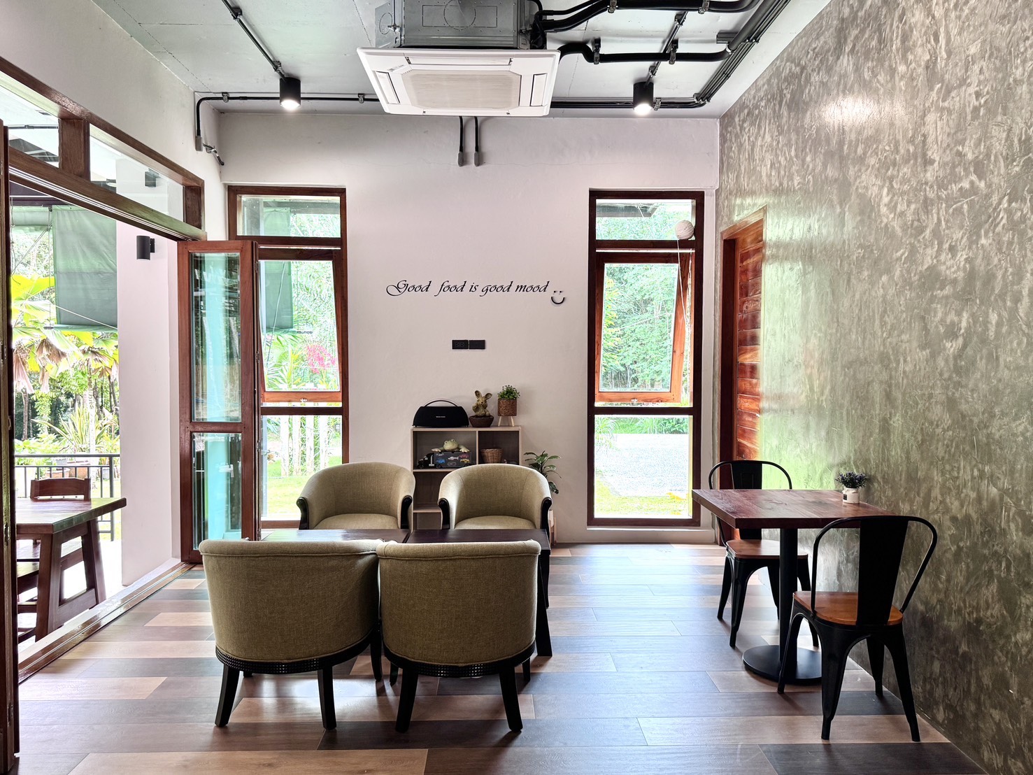 มุมนั่งเล่นภายในร้านคาเฟ่ตกแต่งสไตล์ลอฟท์ มีโซฟาสีเขียวมะกอก โต๊ะไม้ เก้าอี้เหล็กสีดำ ผนังปูนเปลือย และข้อความบนผนัง Good food is good mood