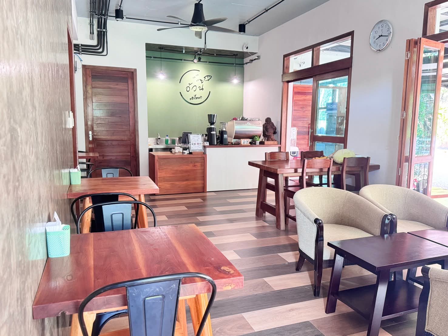 ภายในร้านกาแฟอ้วน บรรยากาศอบอุ่นด้วยโต๊ะเก้าอี้ไม้ เคาน์เตอร์ชงกาแฟ ผนังสีเขียว พร้อมโซฟานั่งสบาย