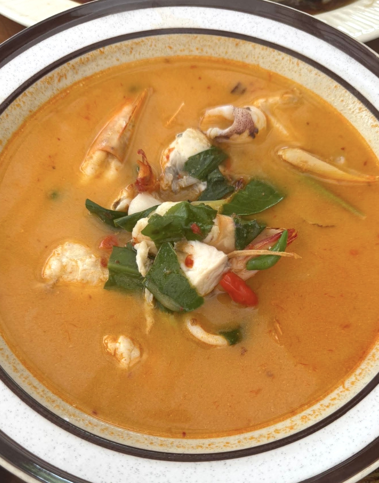 ต้มยำทะเลใส่ปูและกุ้งในชามเซรามิก น้ำซุปสีส้มเข้มข้นตกแต่งด้วยใบโหระพาและพริก