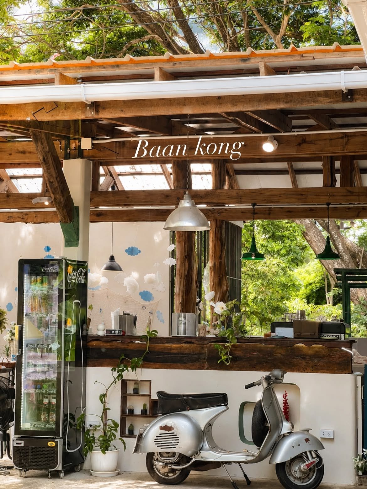 ร้านบ้านกอง คาเฟ่บรรยากาศอบอุ่นสไตล์วินเทจ มีเคาน์เตอร์ไม้เก่า ตู้แช่โค้ก และรถเวสป้าสีเงินจอดหน้าร้าน ใต้หลังคาโครงไม้ร่มรื่น