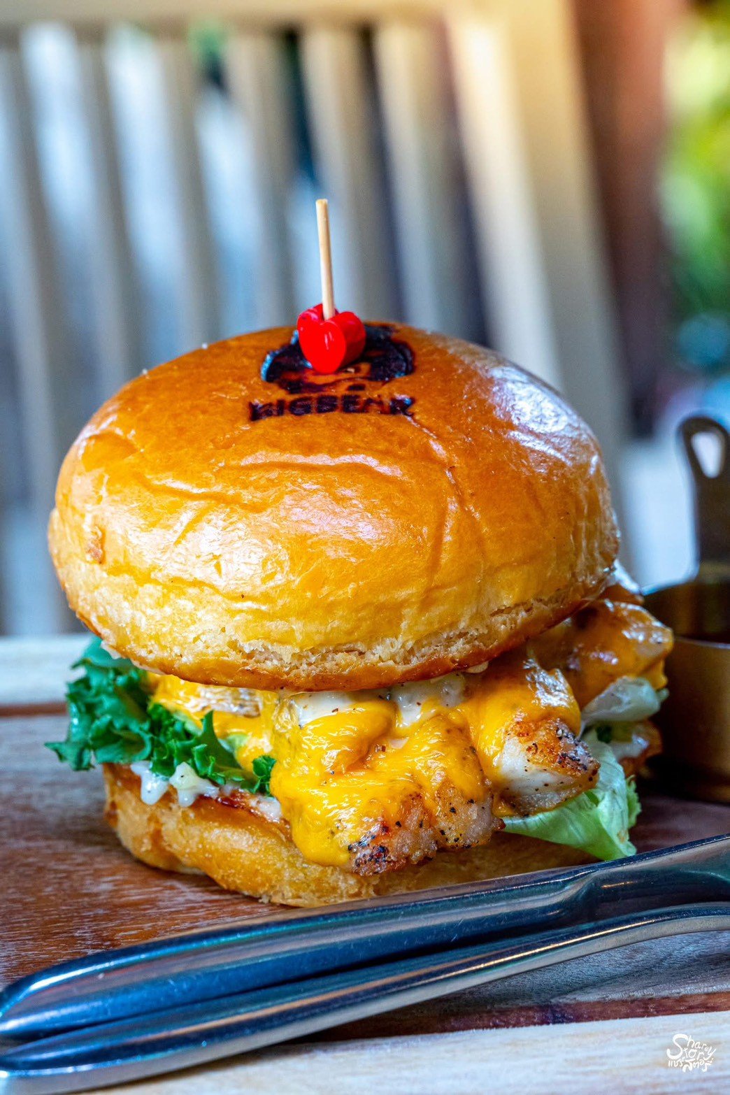 เบอร์เกอร์ไก่ย่างชีสจากร้าน BigBear บนขนมปังบริออชตราร้าน พร้อมผักกรีนและชีสเหลืองละลาย เสิร์ฟบนถาดไม้