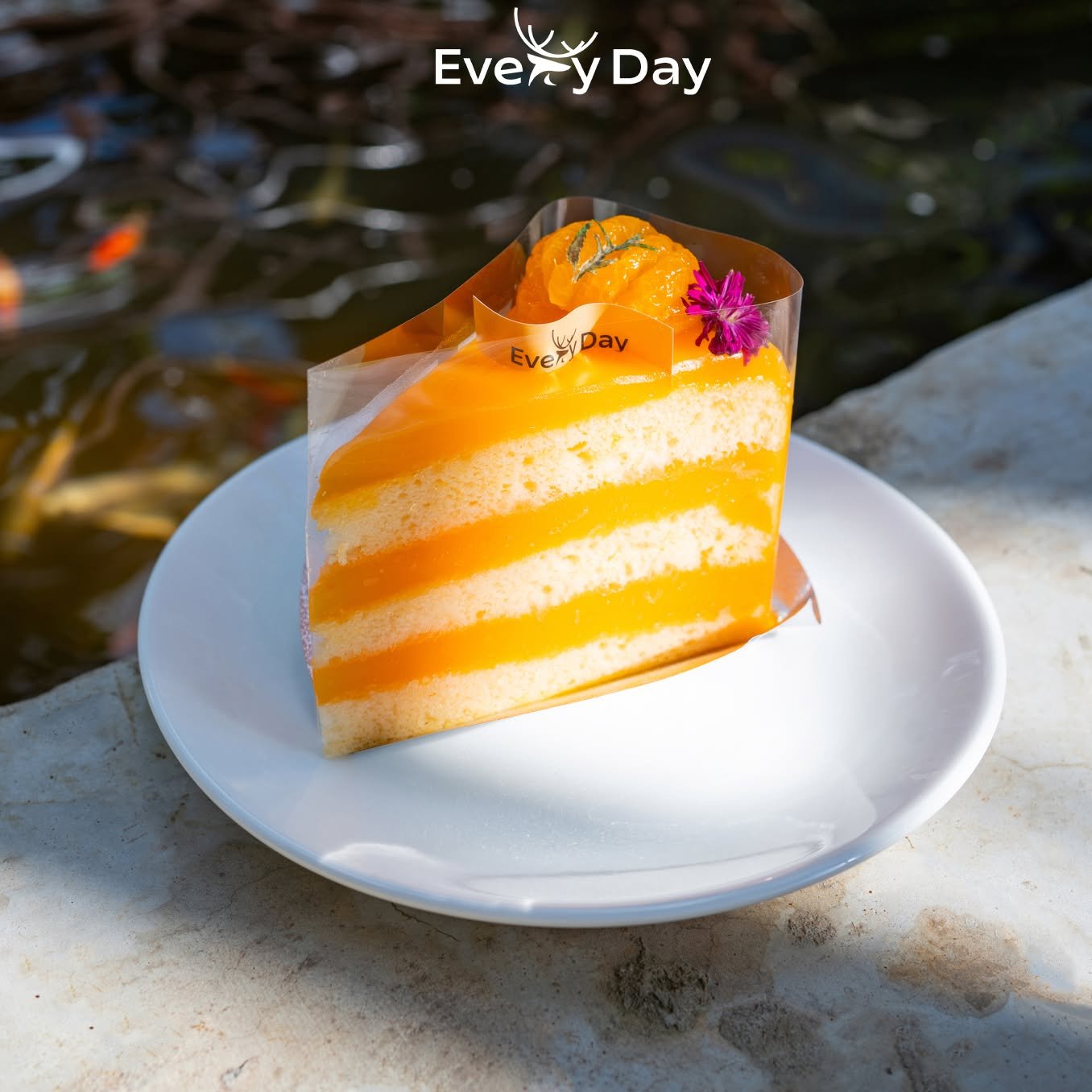 เค้กมะม่วงหลายชั้นตกแต่งด้วยดอกไม้สีชมพูจากร้าน Every Day เสิร์ฟบนจานขาวริมบ่อปลาคาร์ป
