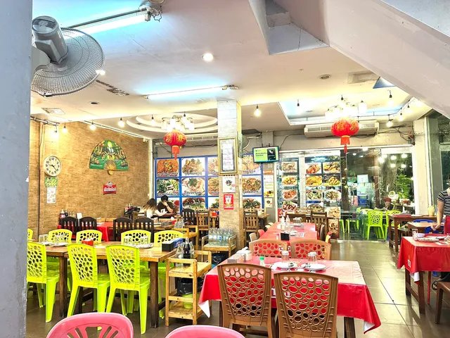 ภายในร้านอาหารไทยที่มีโต๊ะเก้าอี้หลากสีสัน โคมไฟจีนสีแดง พัดลมติดผนัง และเมนูอาหารติดผนังด้านหลัง