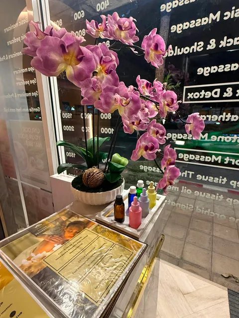 หน้าต่างร้านสปานวดไทยตกแต่งด้วยดอกกล้วยไม้สีชมพูในกระถางขาว พร้อมขวดน้ำมันนวดและใบประกาศนียบัตร มองเห็นเมนูบริการนวดสะท้อนบนกระจก